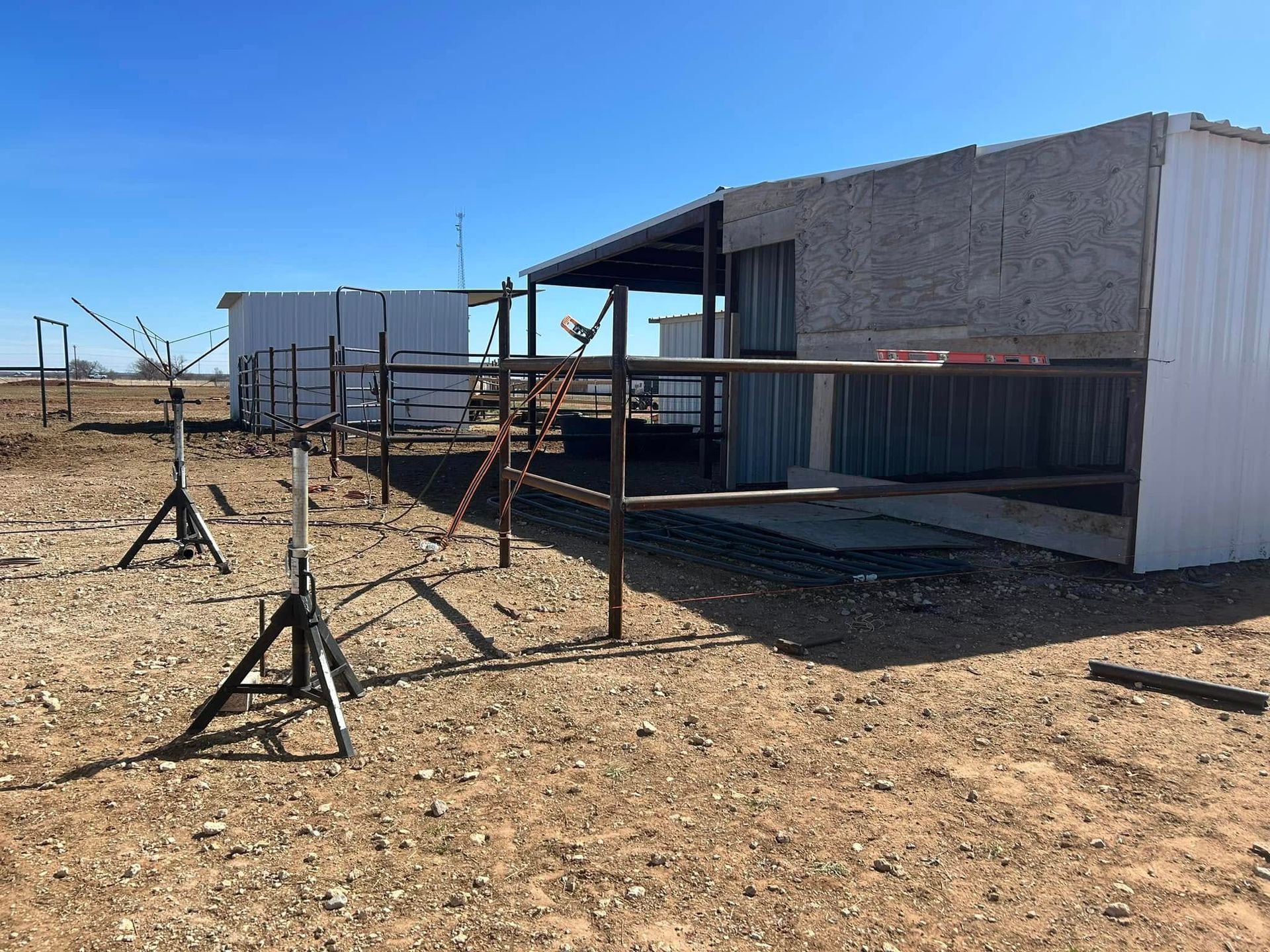 White metal sheds and metal pipe fencing stand on a dry, rocky desert landscape under a clear blue sky.
