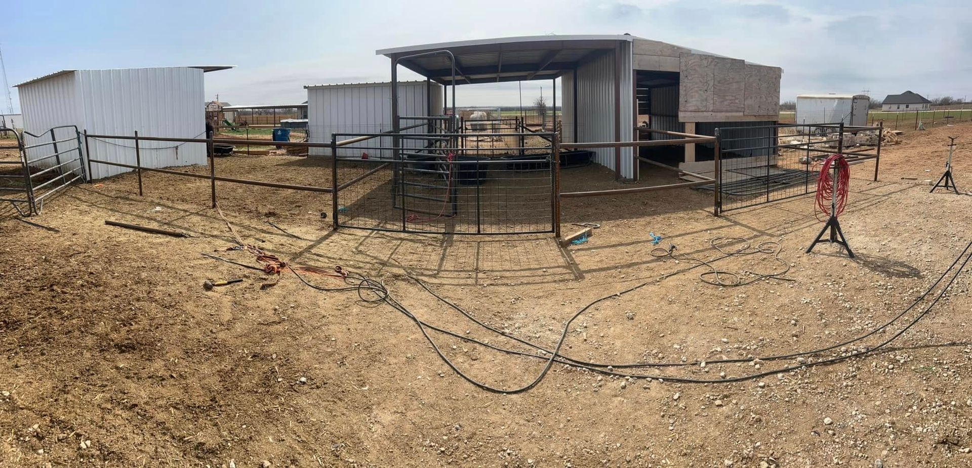 A panoramic view of an outdoor animal enclosure with shelters, fences, and equipment on dry, dusty ground under a clear sky.