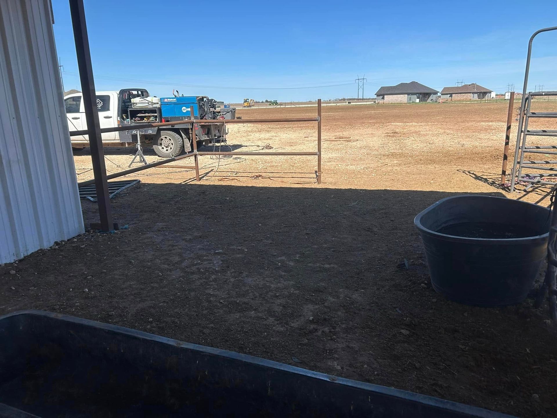 A truck with a welding machine parked in a dirt field near a corrugated metal structure and livestock water troughs.