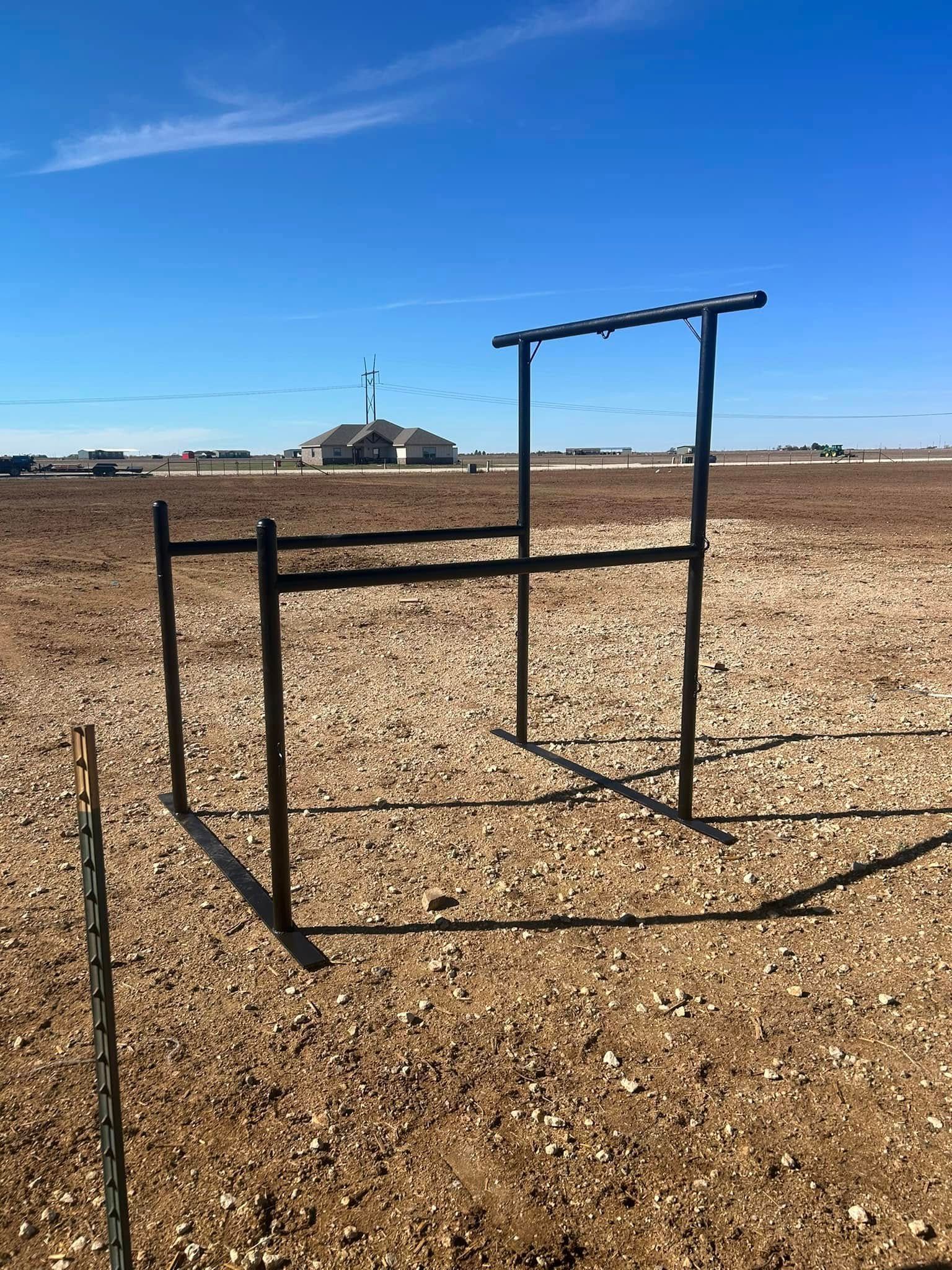 A black metal frame structure stands in a flat, rocky field under a clear blue sky, with a house visible in the distance.