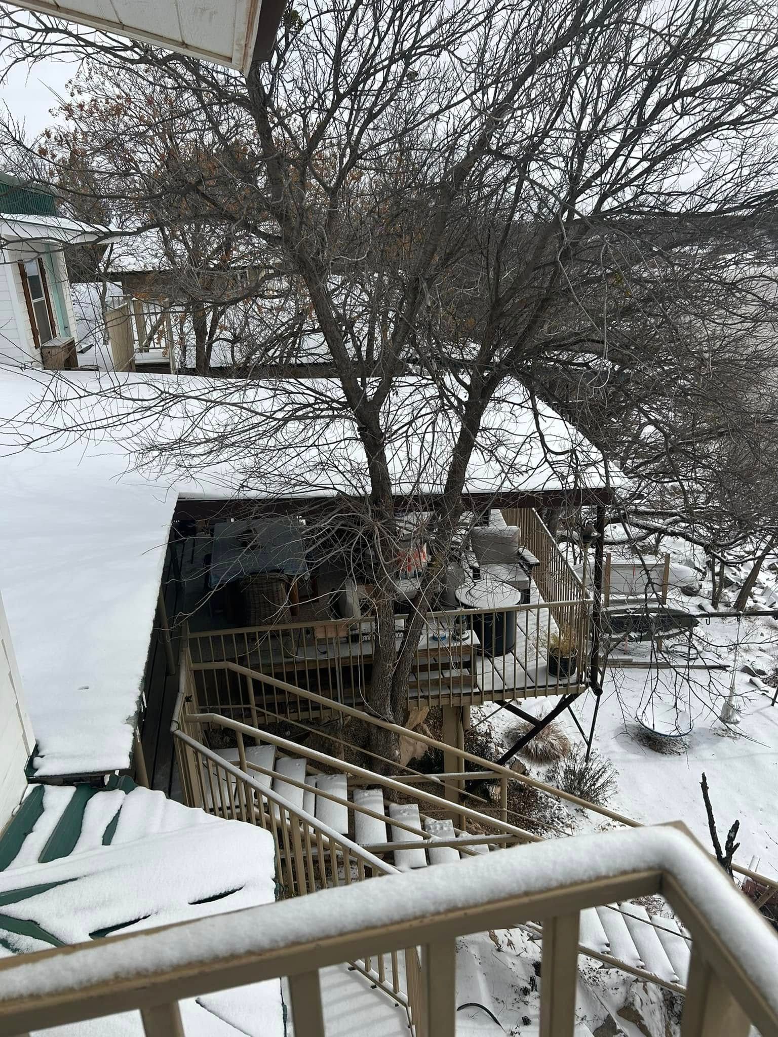 Snow-covered wooden porch and staircase descending toward a tree and a snow-dusted structure in a winter landscape.