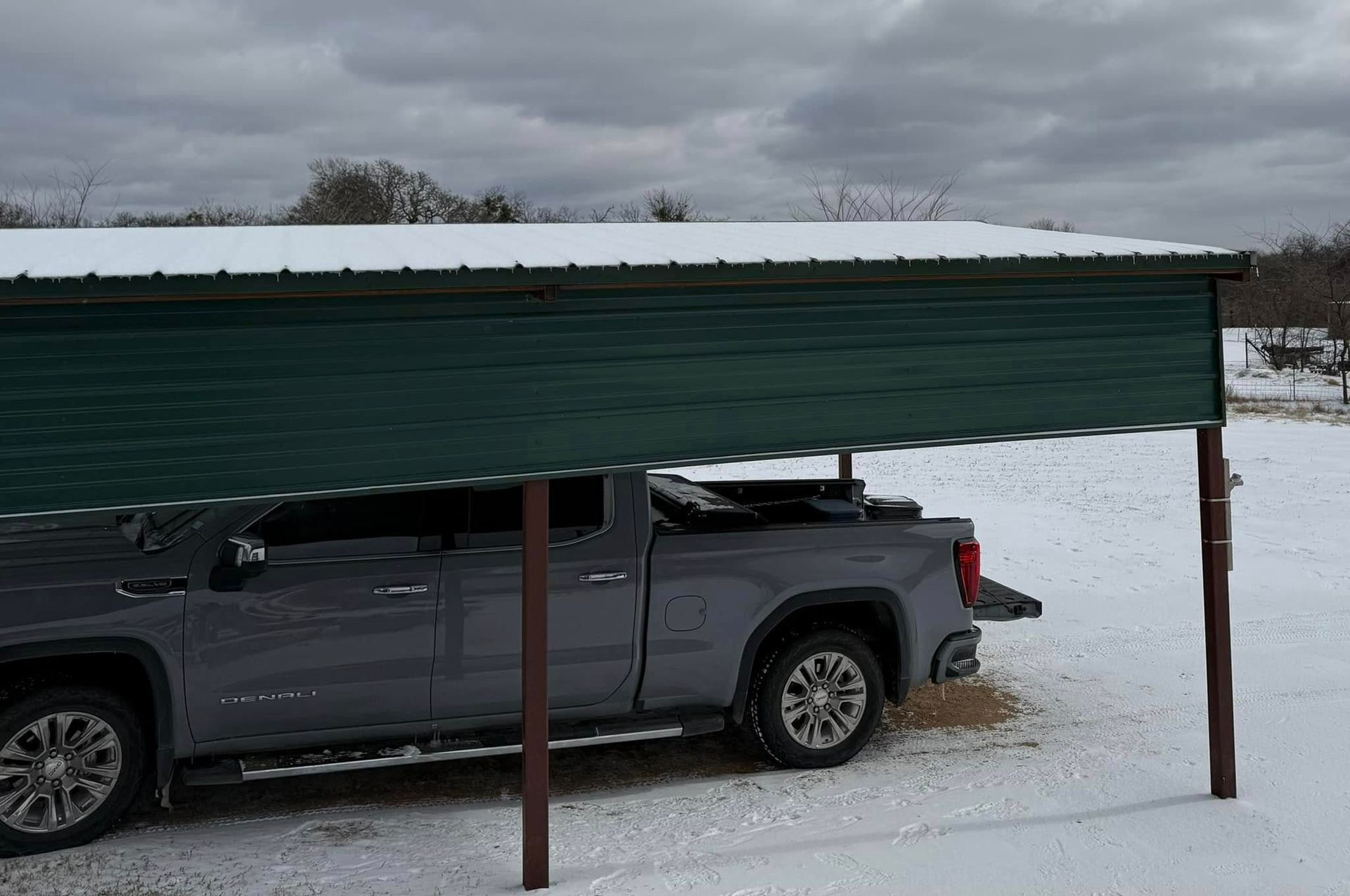 A gray pickup truck is parked under a green metal carport in a snow-covered field under a cloudy winter sky.