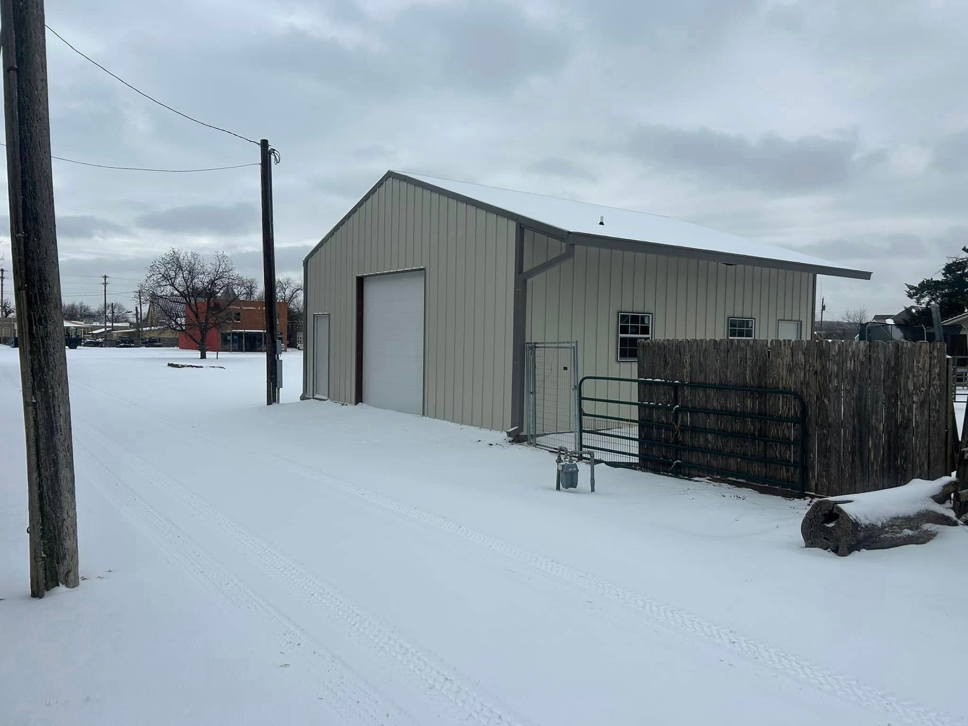 A metal outbuilding with a large garage door stands in a snow-covered yard under a cloudy sky.