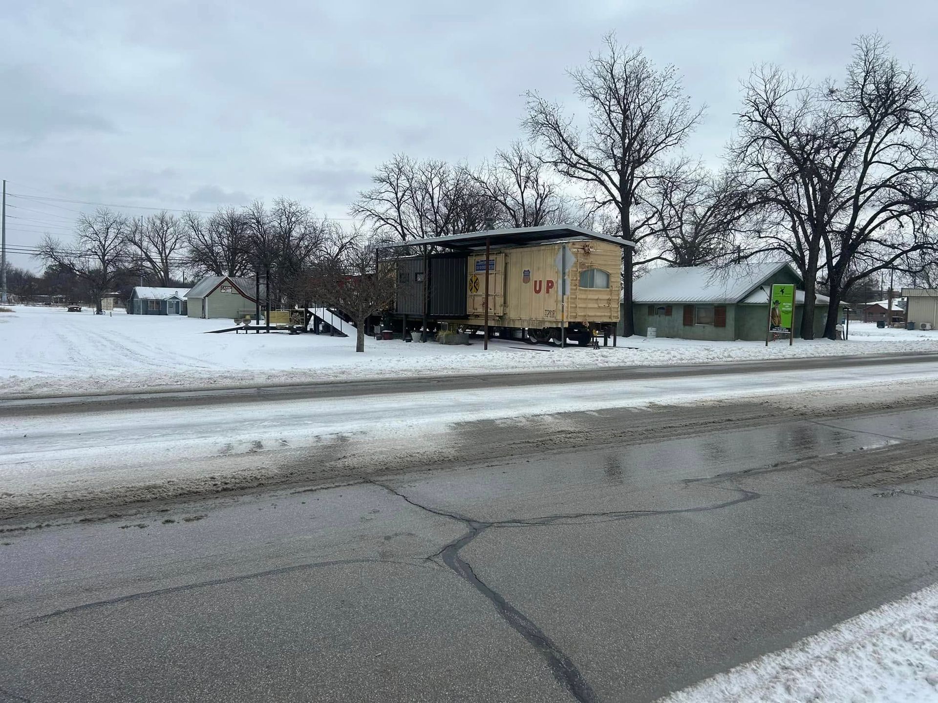 A tan, aged trailer stands on a snow-covered field next to a small house under a cloudy sky near a paved road.