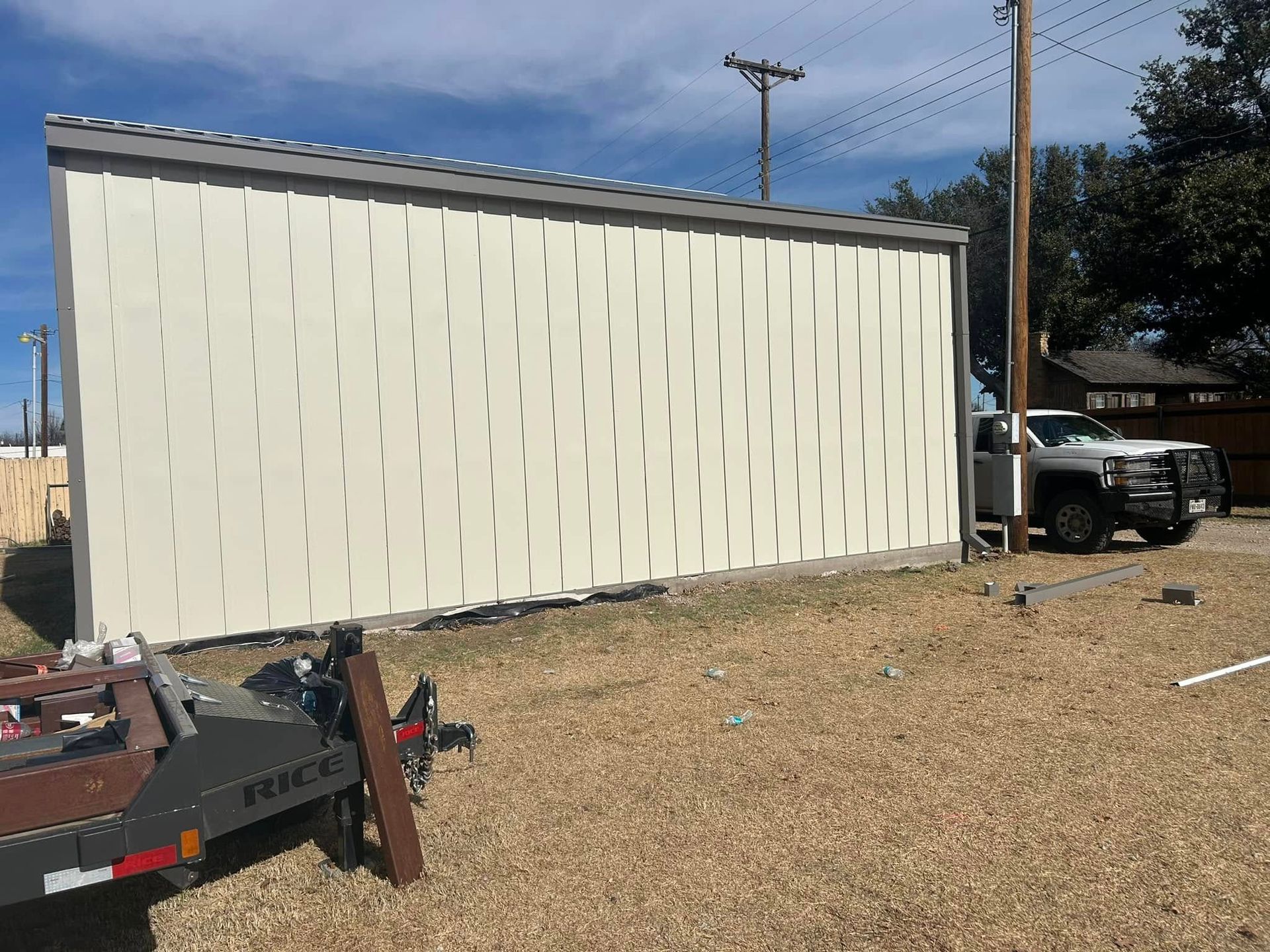 A long, beige corrugated metal wall stands on a gravel lot with a silver pickup truck parked to the right.