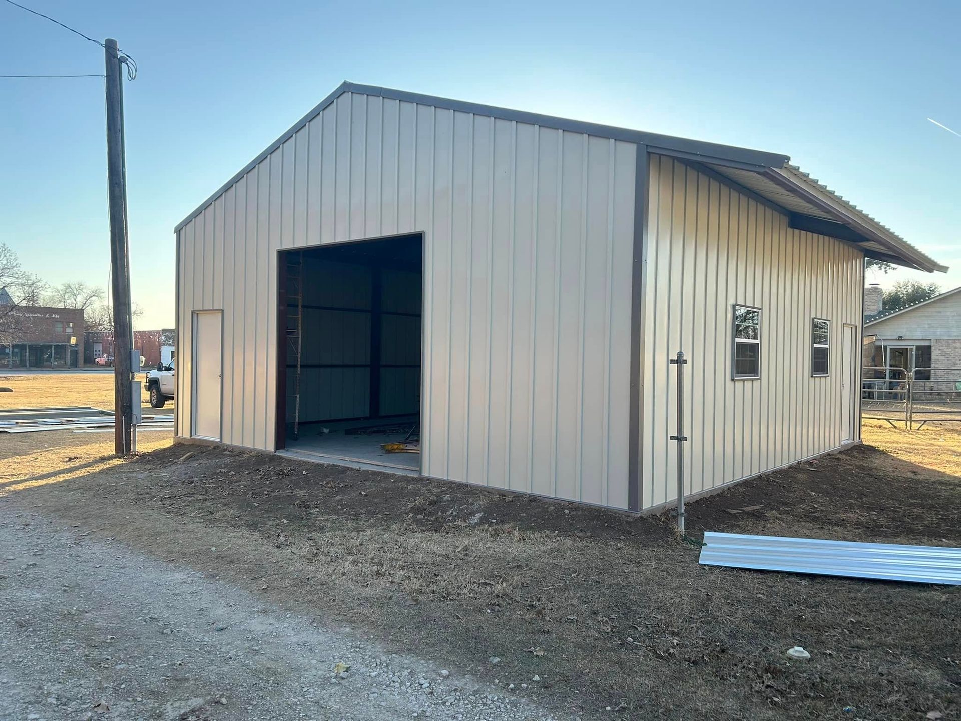 A beige metal workshop building under construction with a large open bay door and two windows on a gravel lot.