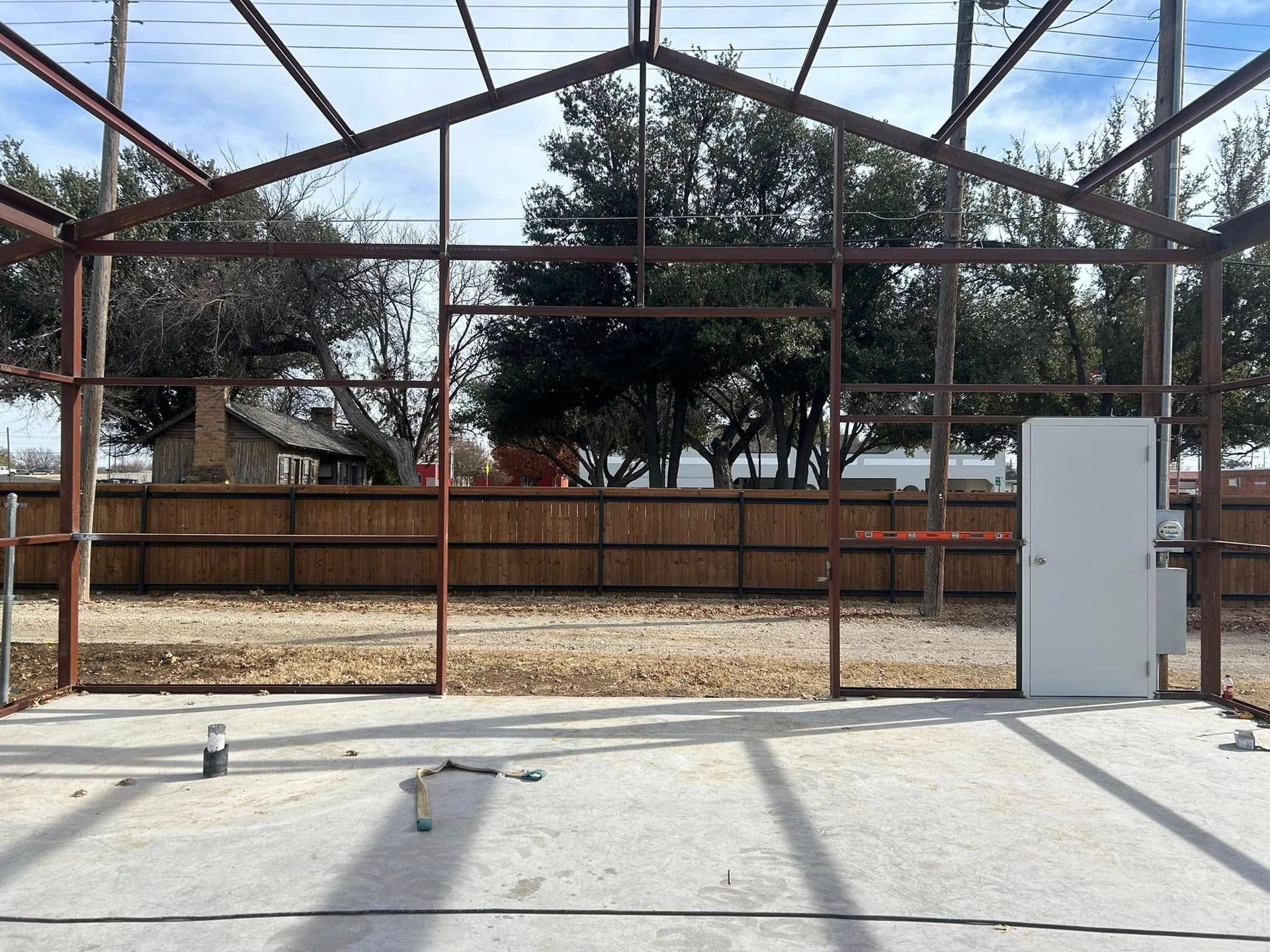 A metal frame structure under construction on a concrete slab, with a lone white door installed against a wood fence.