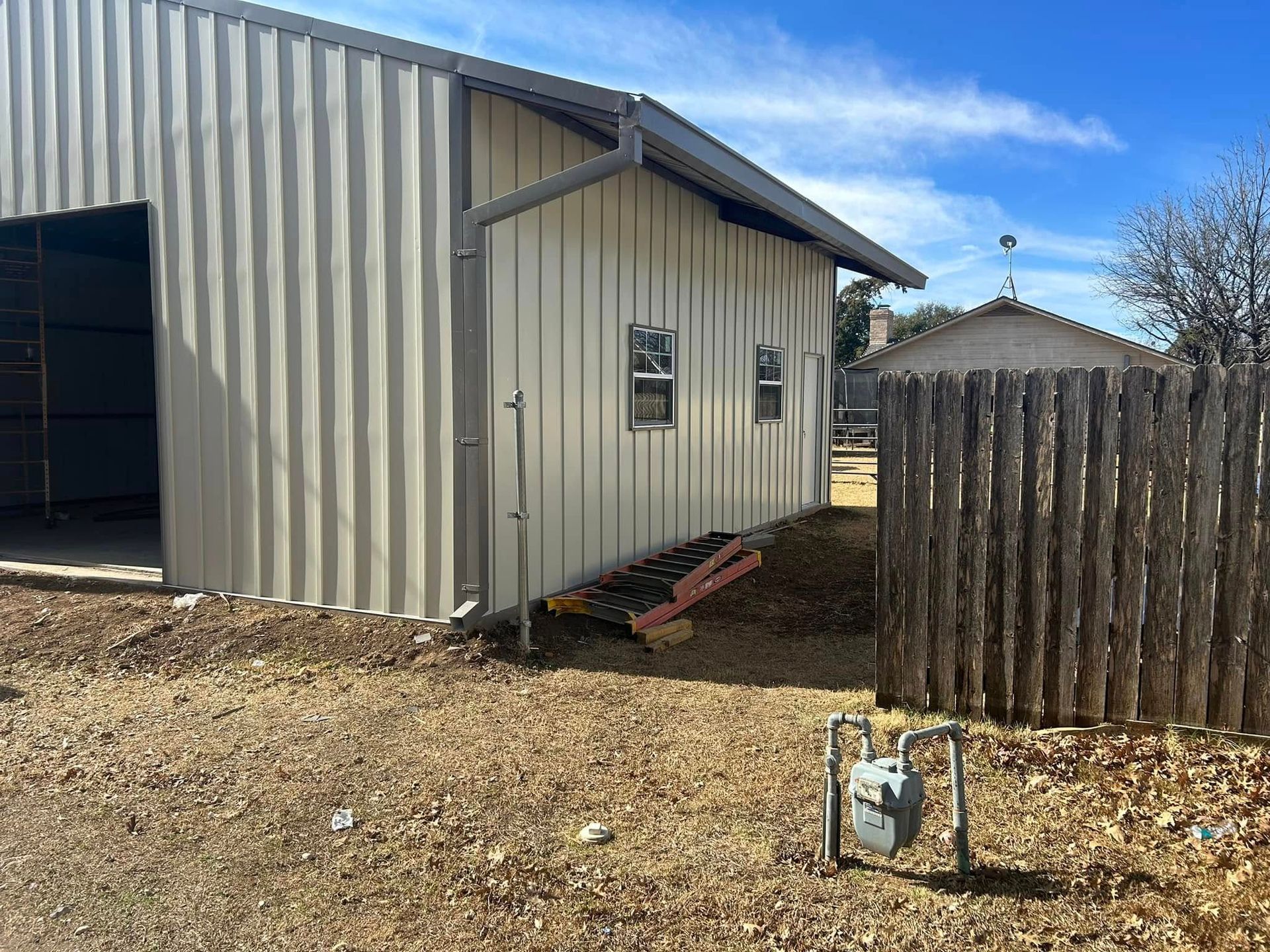 A beige metal building exterior with a downspout, two windows, and a wooden fence, seen on a sunny day.