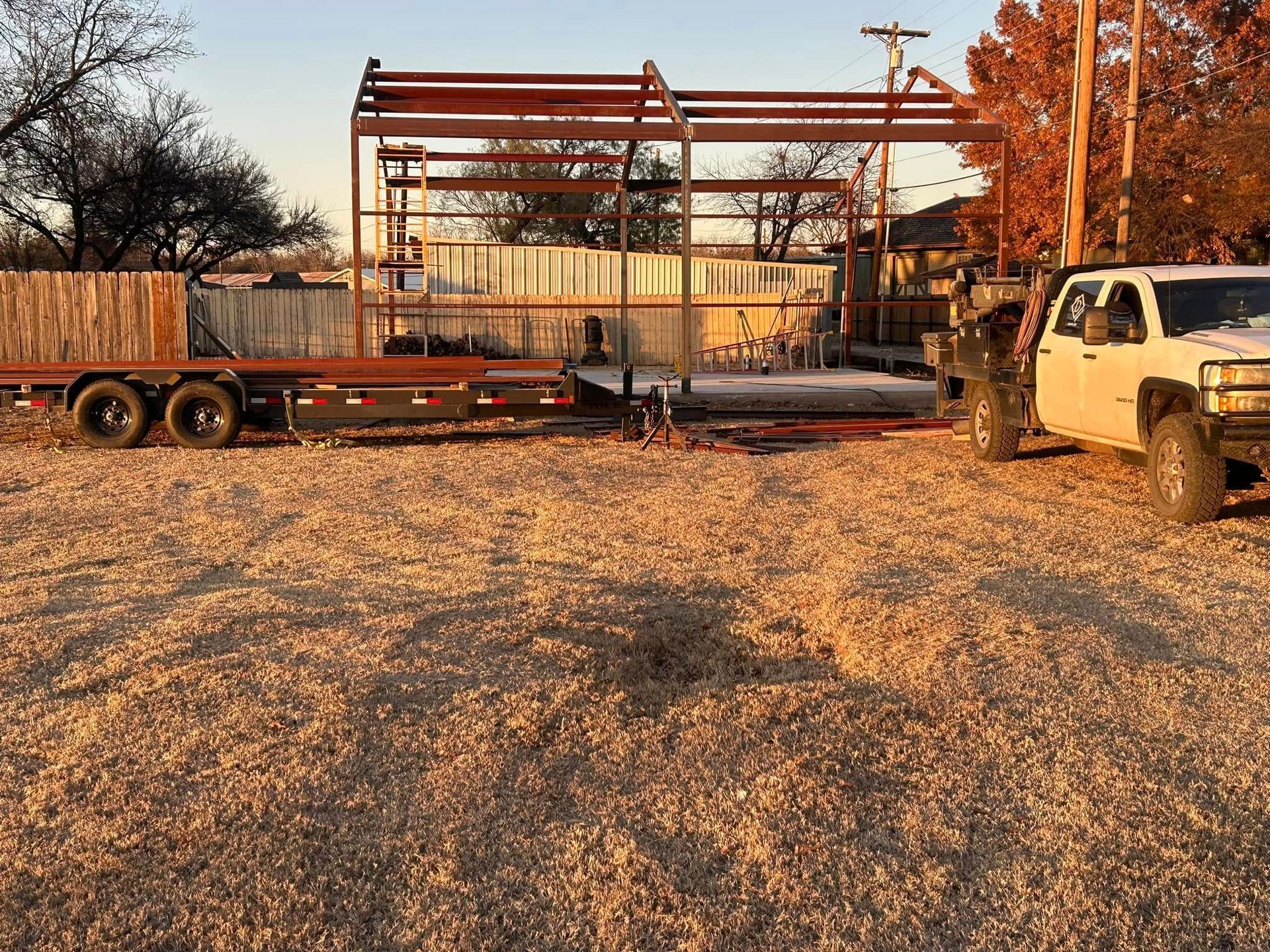 A white pickup truck parked next to a flatbed trailer in front of a partially constructed metal building at sunset.