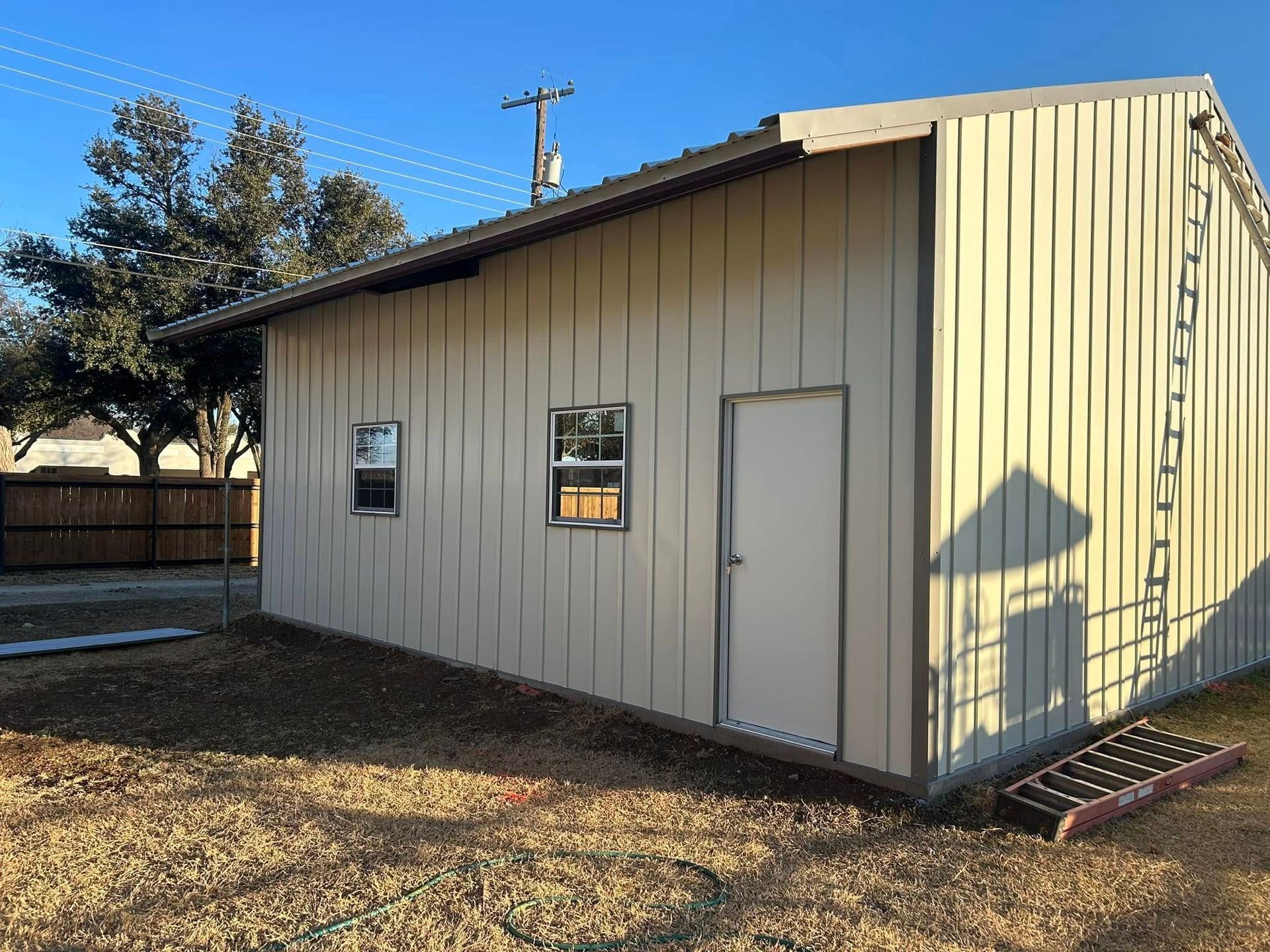 A light-tan metal utility shed with two windows and a white door, set on a gravel lot under a clear blue sky.