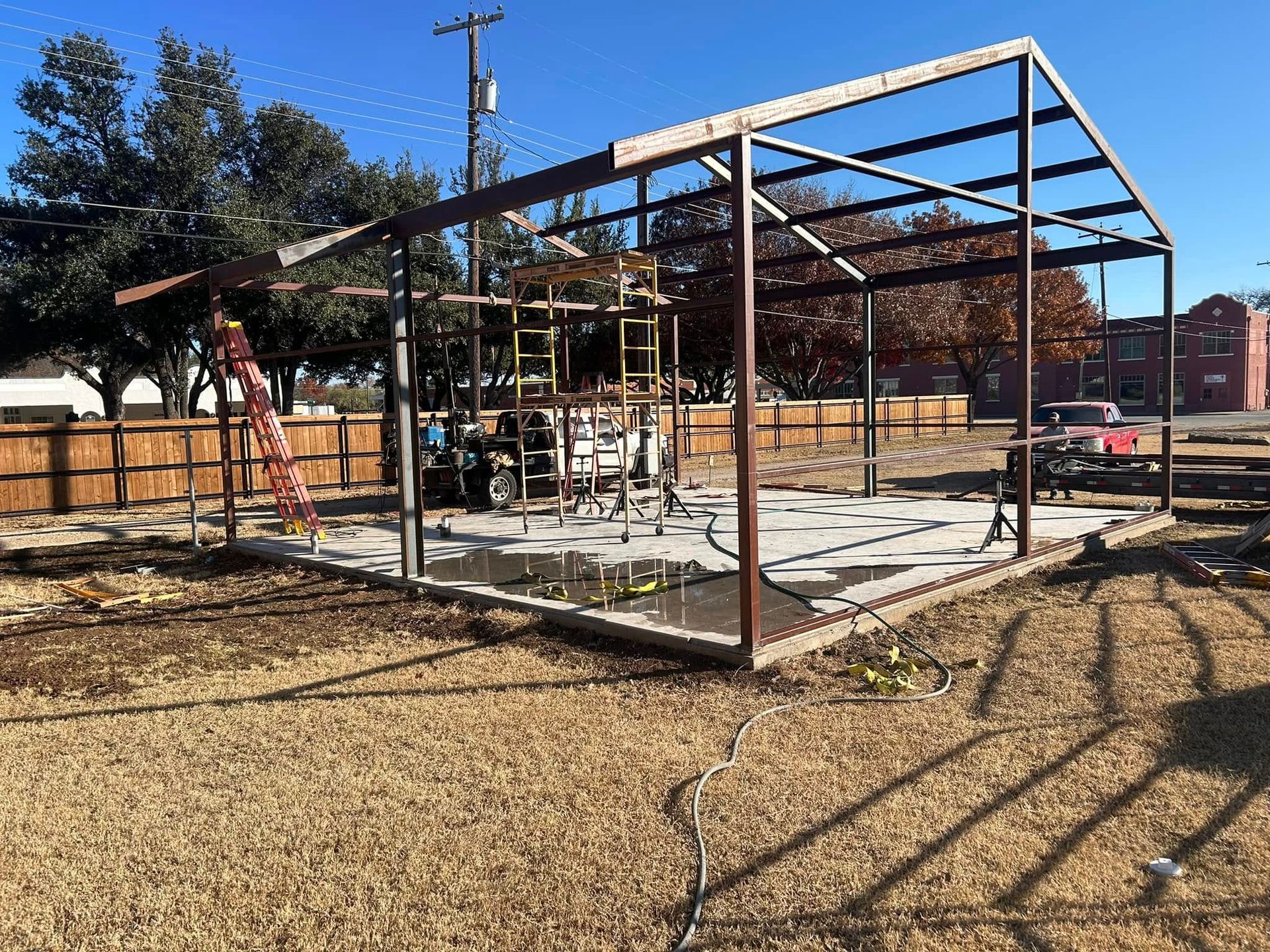 A metal frame structure for a building under construction on a concrete foundation in a grassy lot on a sunny day.