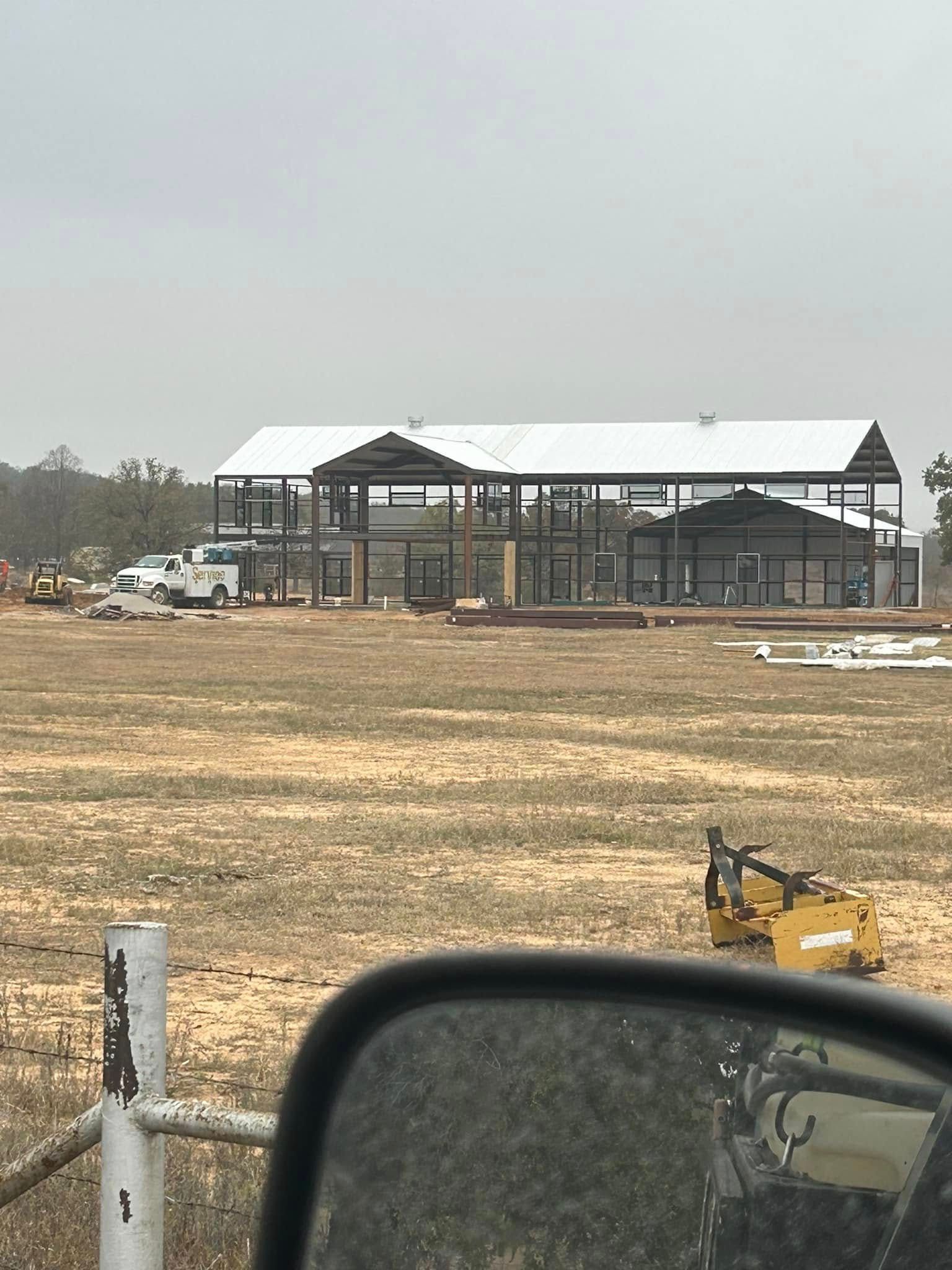 A metal-framed building under construction on a field, viewed from a car with part of the side mirror in the foreground.