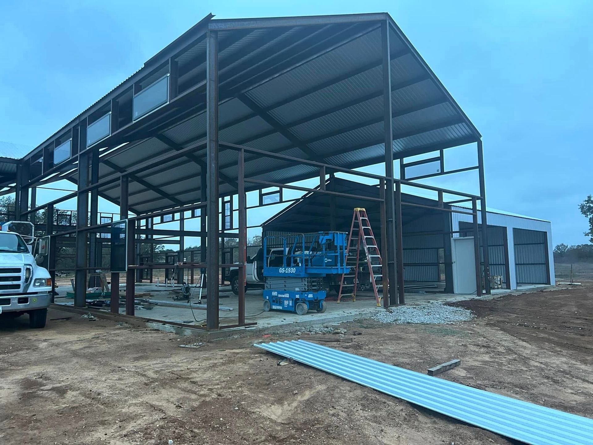 A large metal-framed building under construction with a blue scissor lift inside and a truck parked on the left.