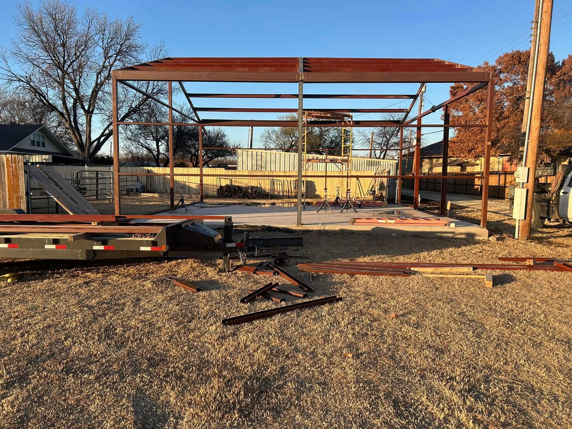 A partially constructed steel building frame stands on a gravel lot under a clear sky, with a flatbed trailer nearby.