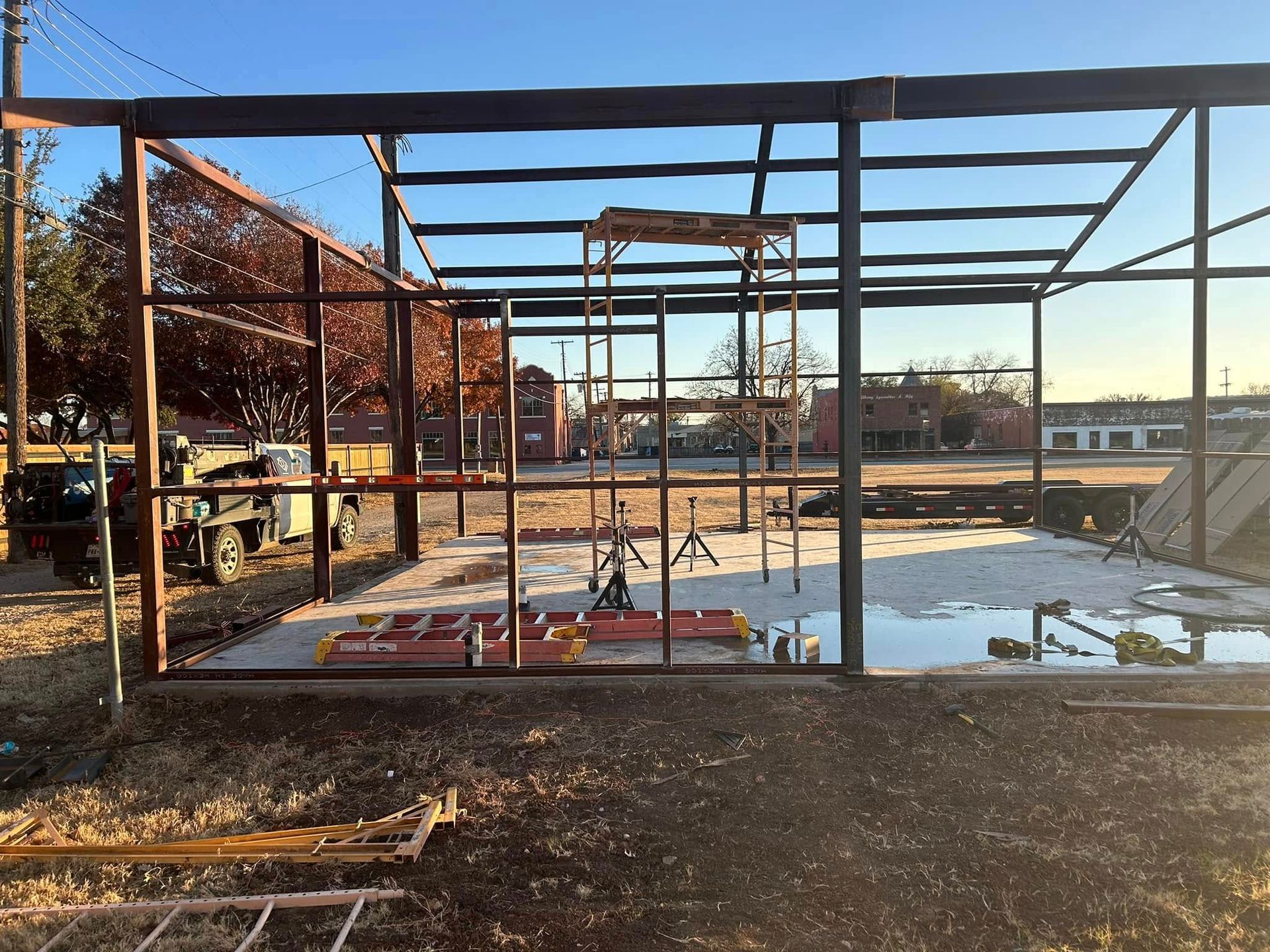 Steel frame structure of a building under construction on a dirt lot, featuring scaffolding and scattered materials.
