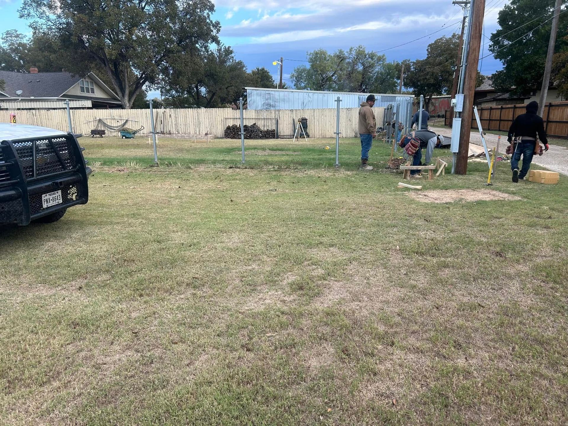 Workers install a chain-link fence on a grassy lot next to a parked black vehicle.