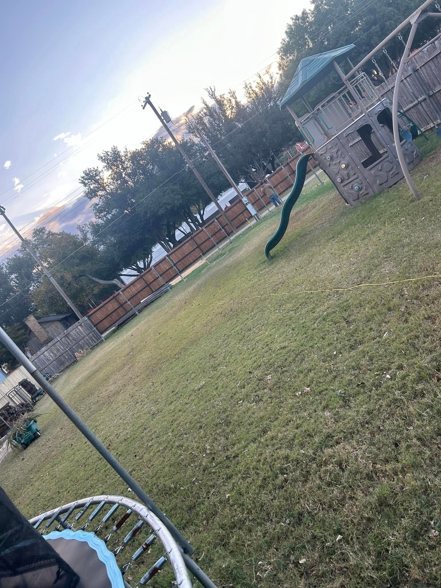 A grassy backyard featuring a wooden play set with a green slide and the edge of a trampoline in the foreground.