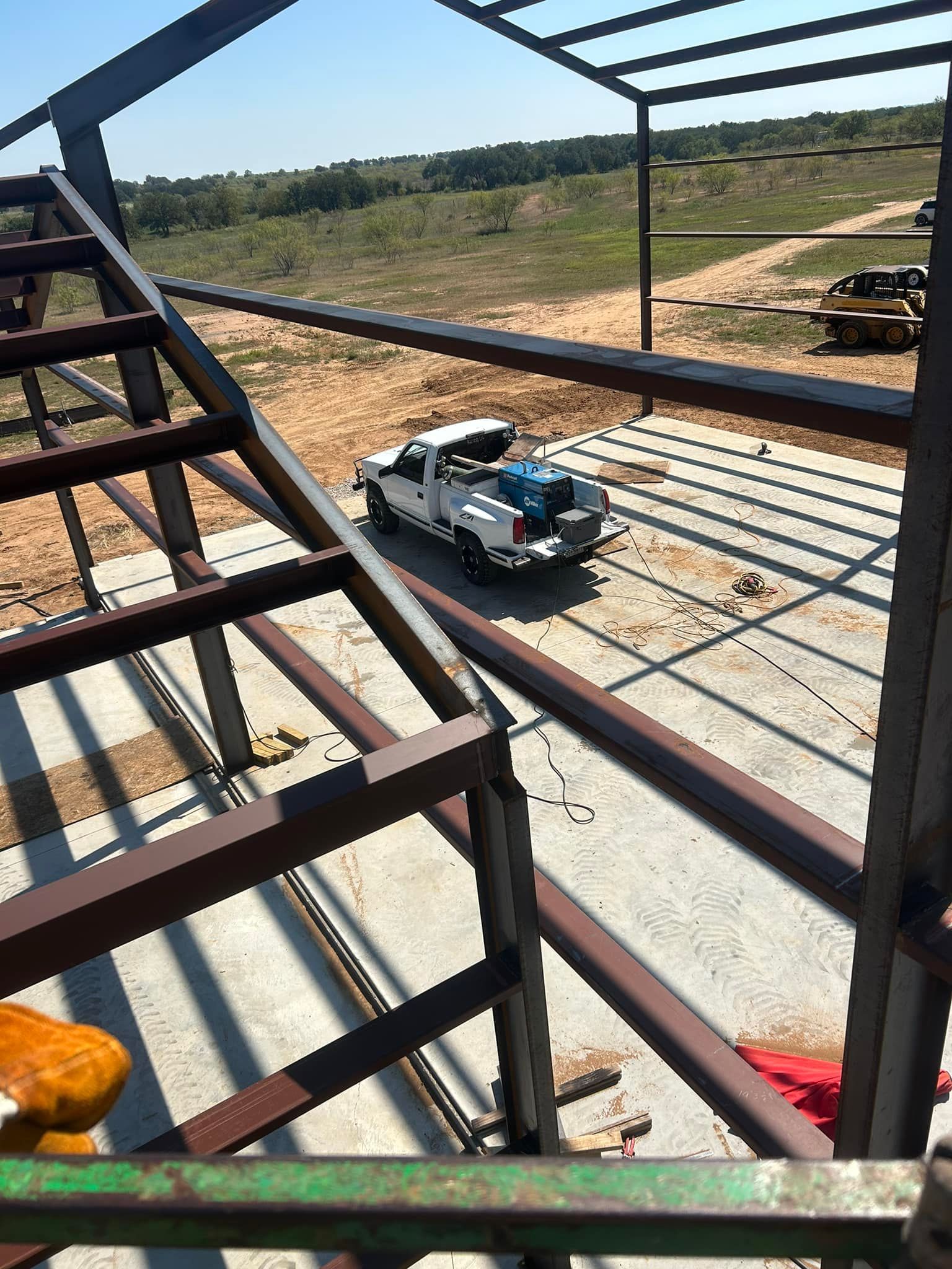 A white truck parked inside the metal frame of a building under construction, overlooking a grassy, rural landscape.