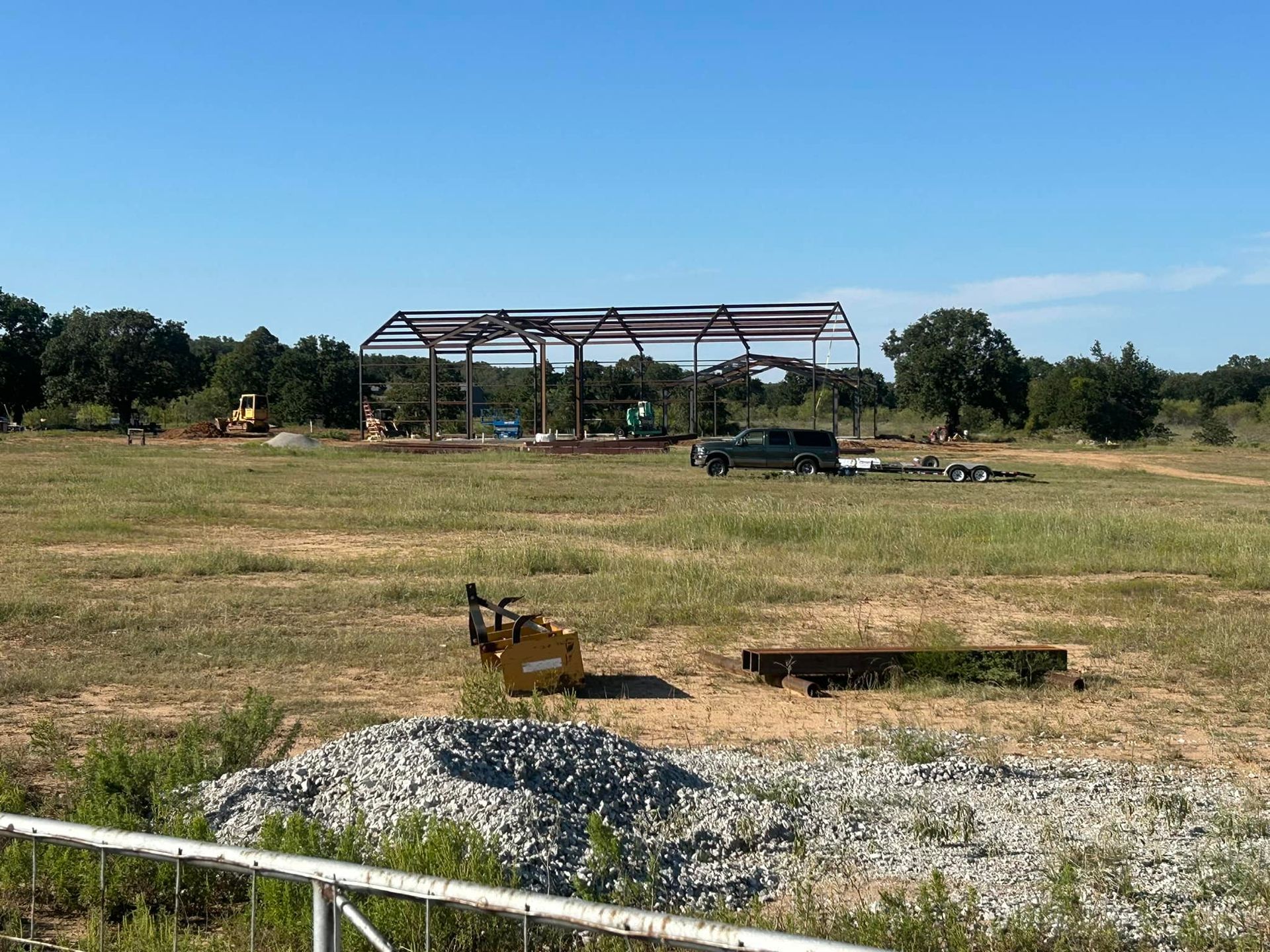 A metal building frame stands in an open, grassy field under a clear blue sky, with construction equipment nearby.