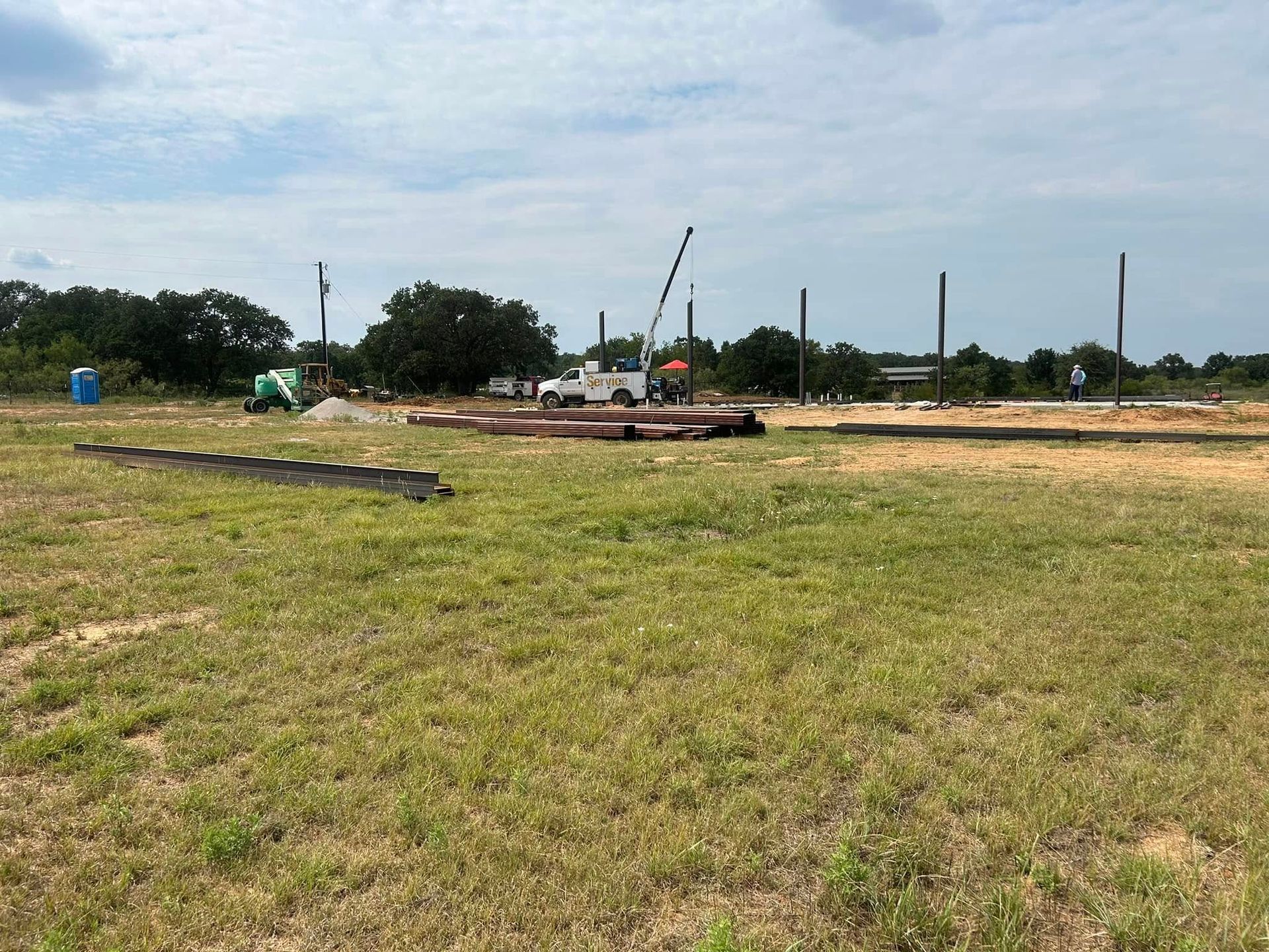 Construction site with vertical steel beams and a white crane truck in an open, grassy field under a cloudy sky.
