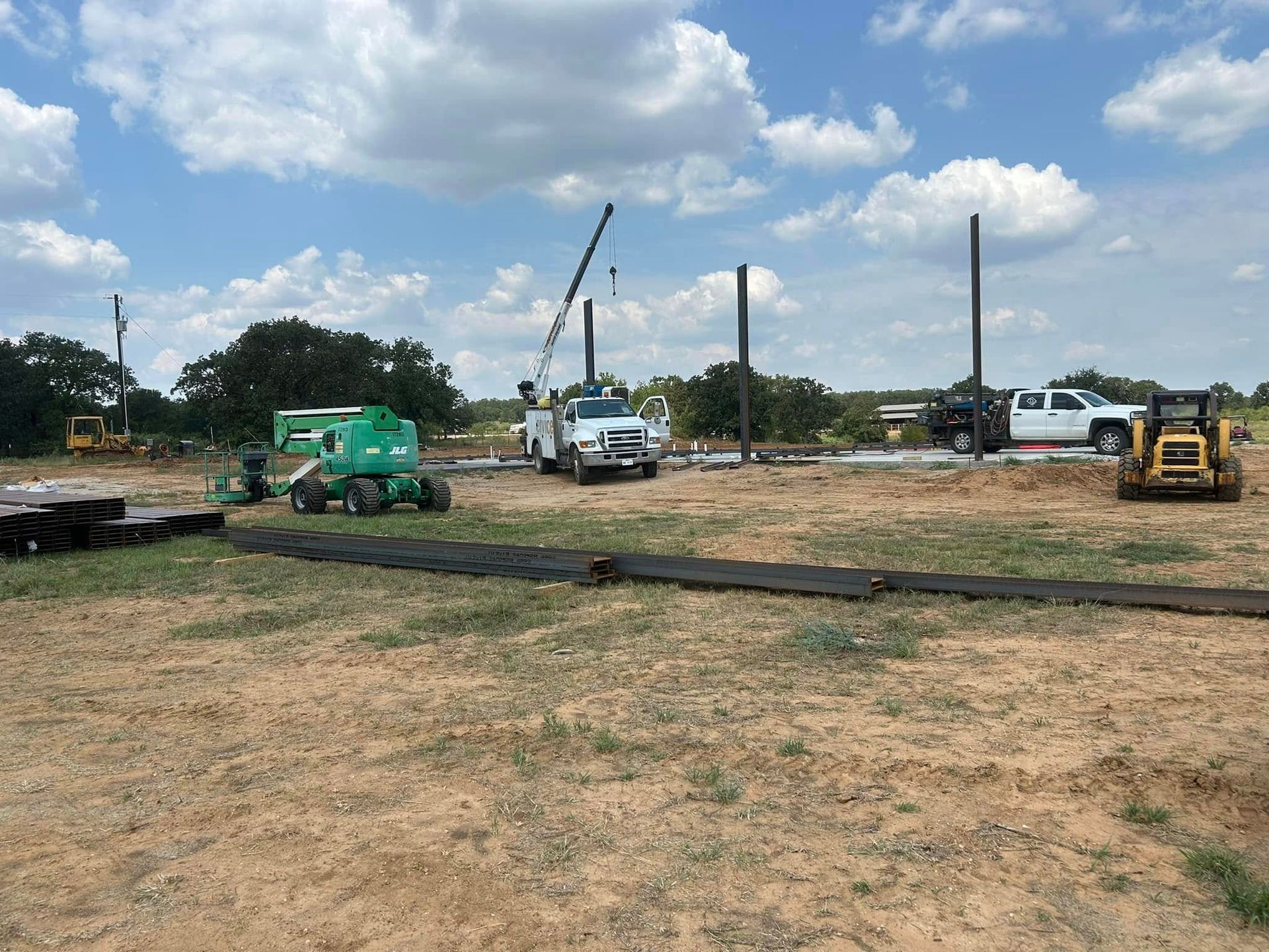 A construction site featuring a crane truck, a green manlift, and a yellow skid-steer on a dirt field with blue skies.