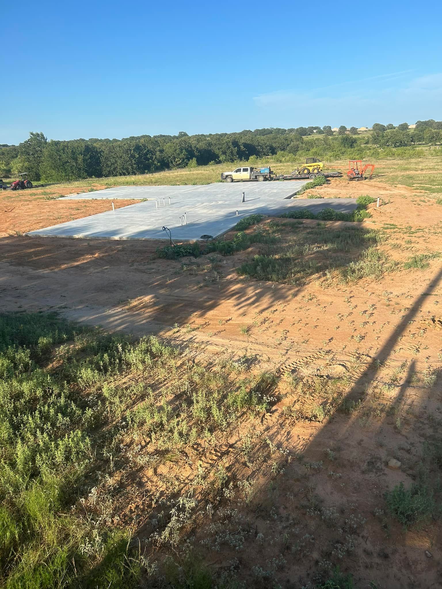 A rectangular concrete foundation slab sits on a dirt lot in a rural, sunny setting with trees in the background.