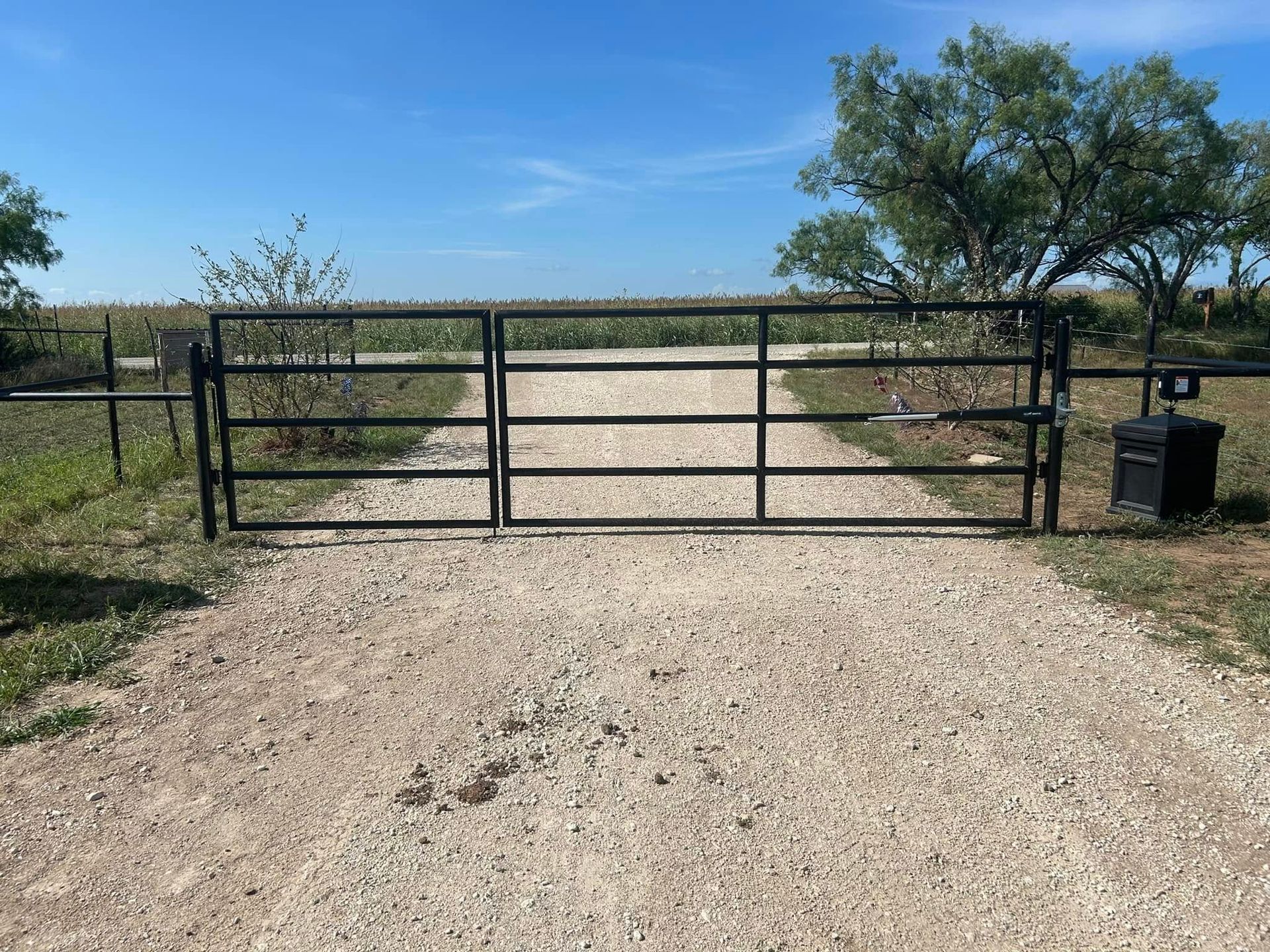 A double black metal farm gate stands closed on a gravel path, leading to a field under a clear blue sky.
