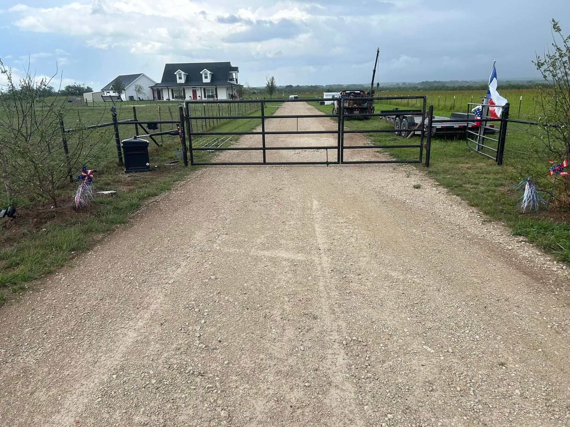 A closed metal gate blocks a gravel driveway leading to a rural house under a cloudy sky.