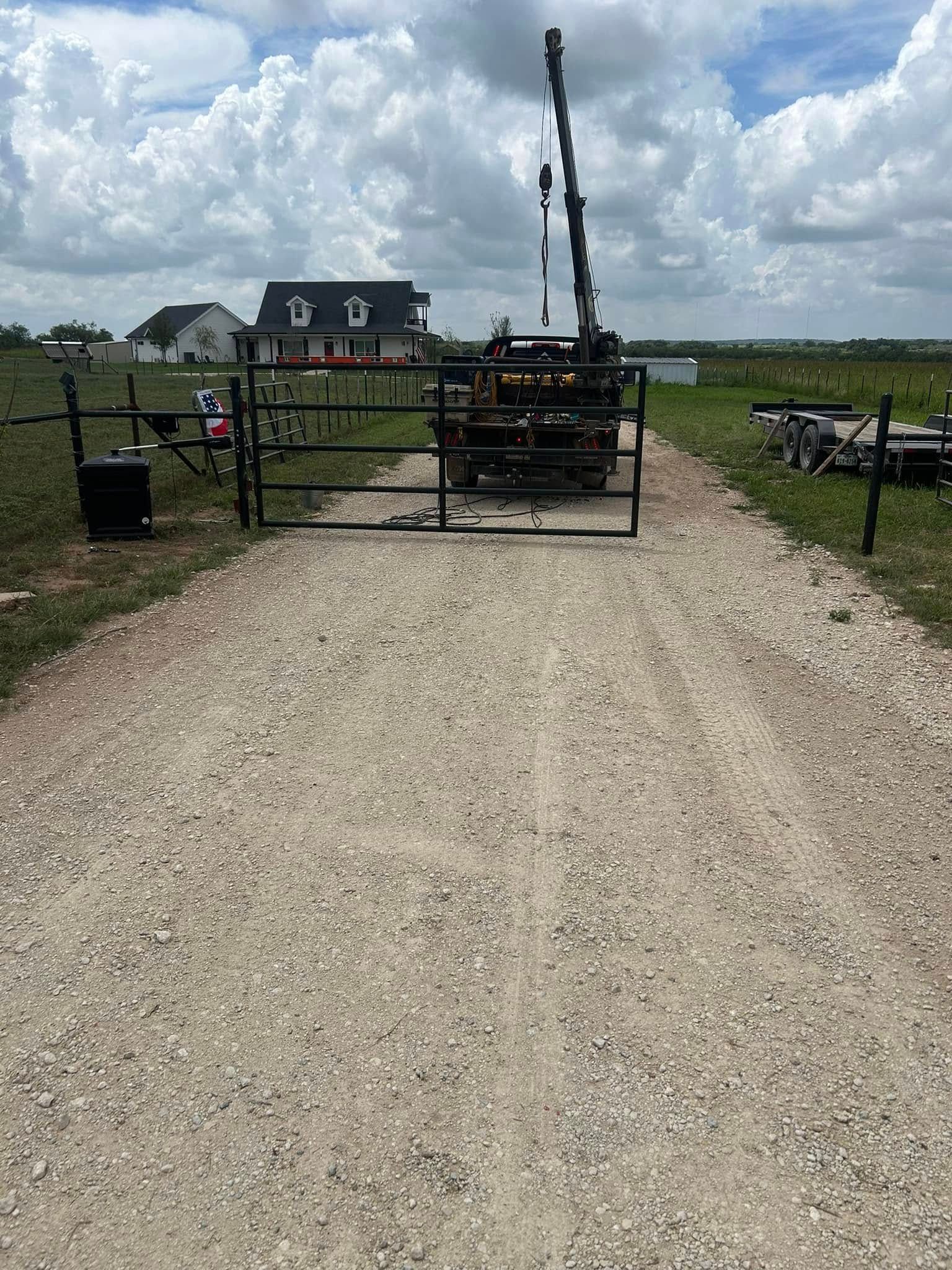 A crane mounted on a truck sits behind a metal gate on a gravel driveway, with a house visible in the background.