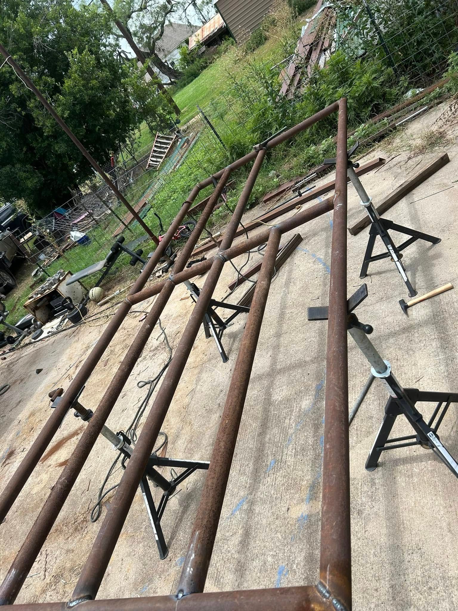 A metal gate frame made of dark brown pipes sits on three adjustable stands on a gravel driveway outdoors.