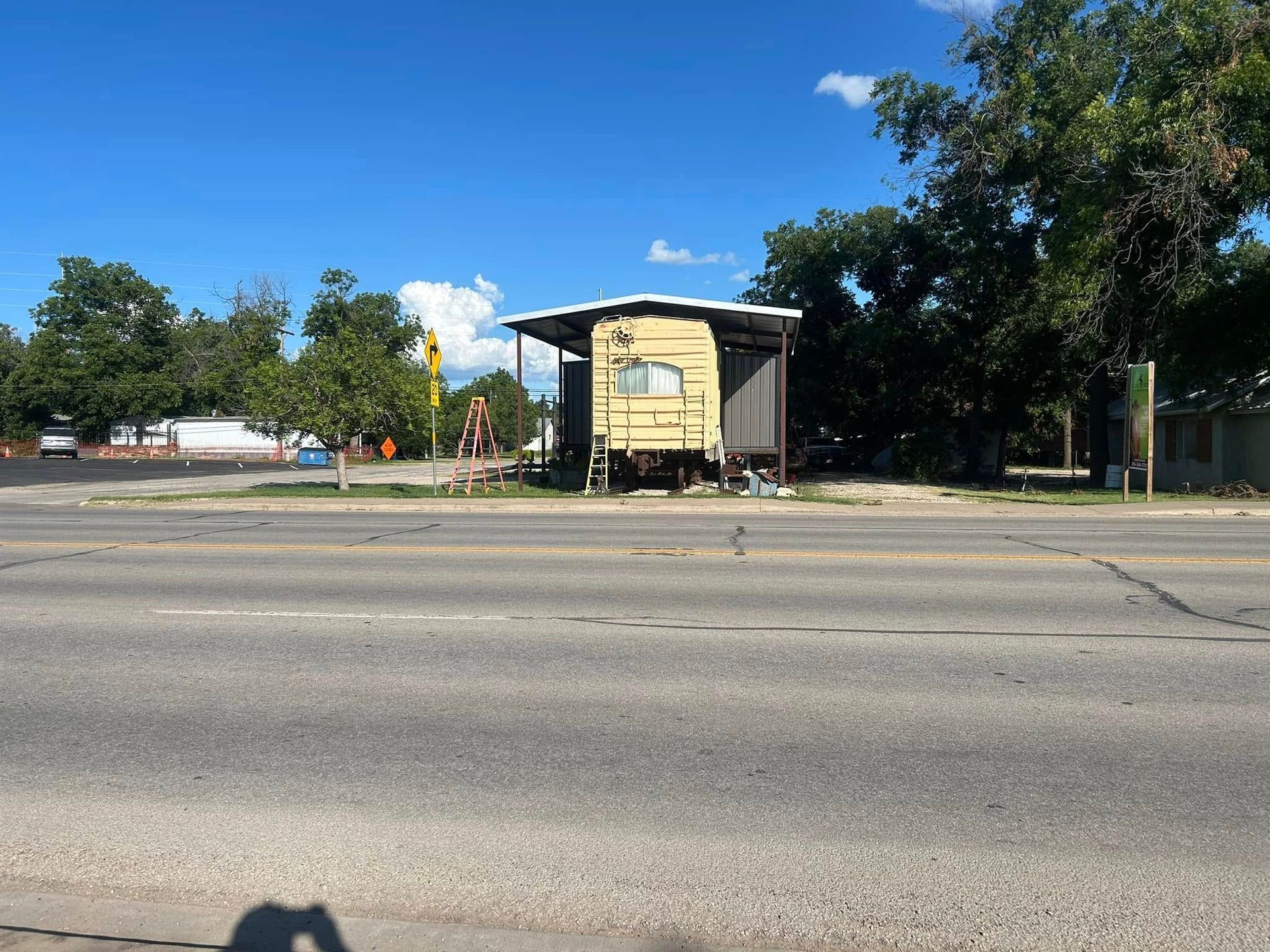 A worn, yellow trailer with a roof extension sits beside a paved road under a clear blue sky, with trees in the background.