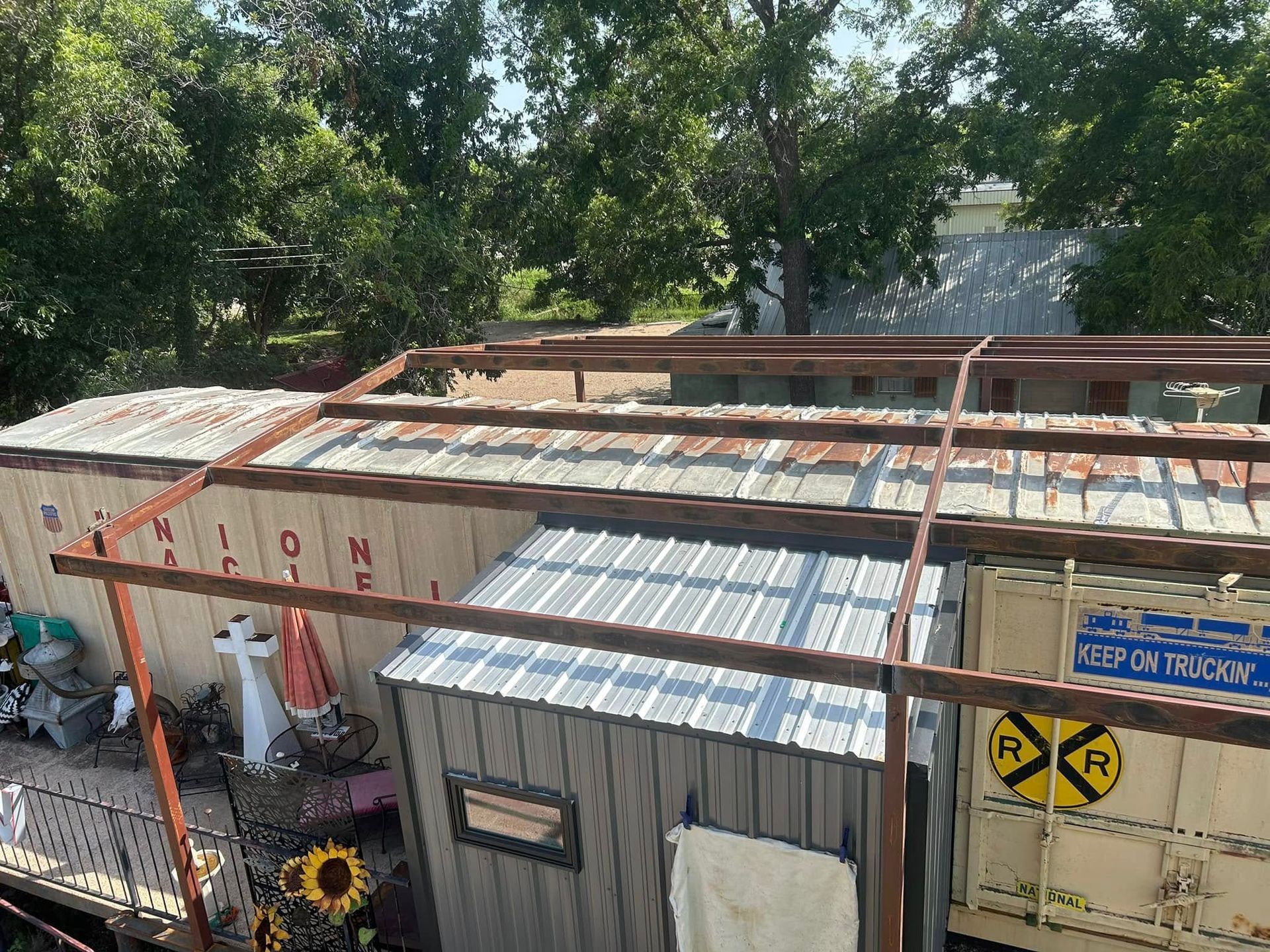 A metal frame structure is built over a storage container and a small shed in a tree-filled outdoor area.