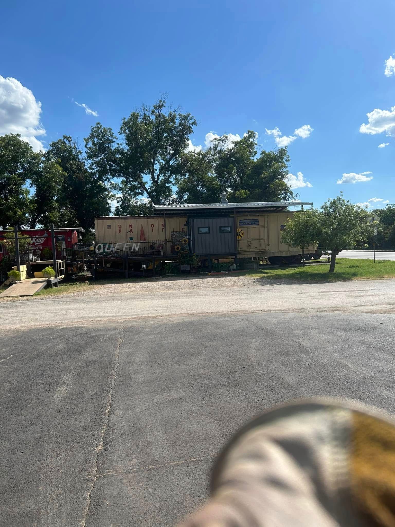 A weathered, tan-colored train caboose sits in a gravel lot under a bright, clear blue sky with scattered trees.