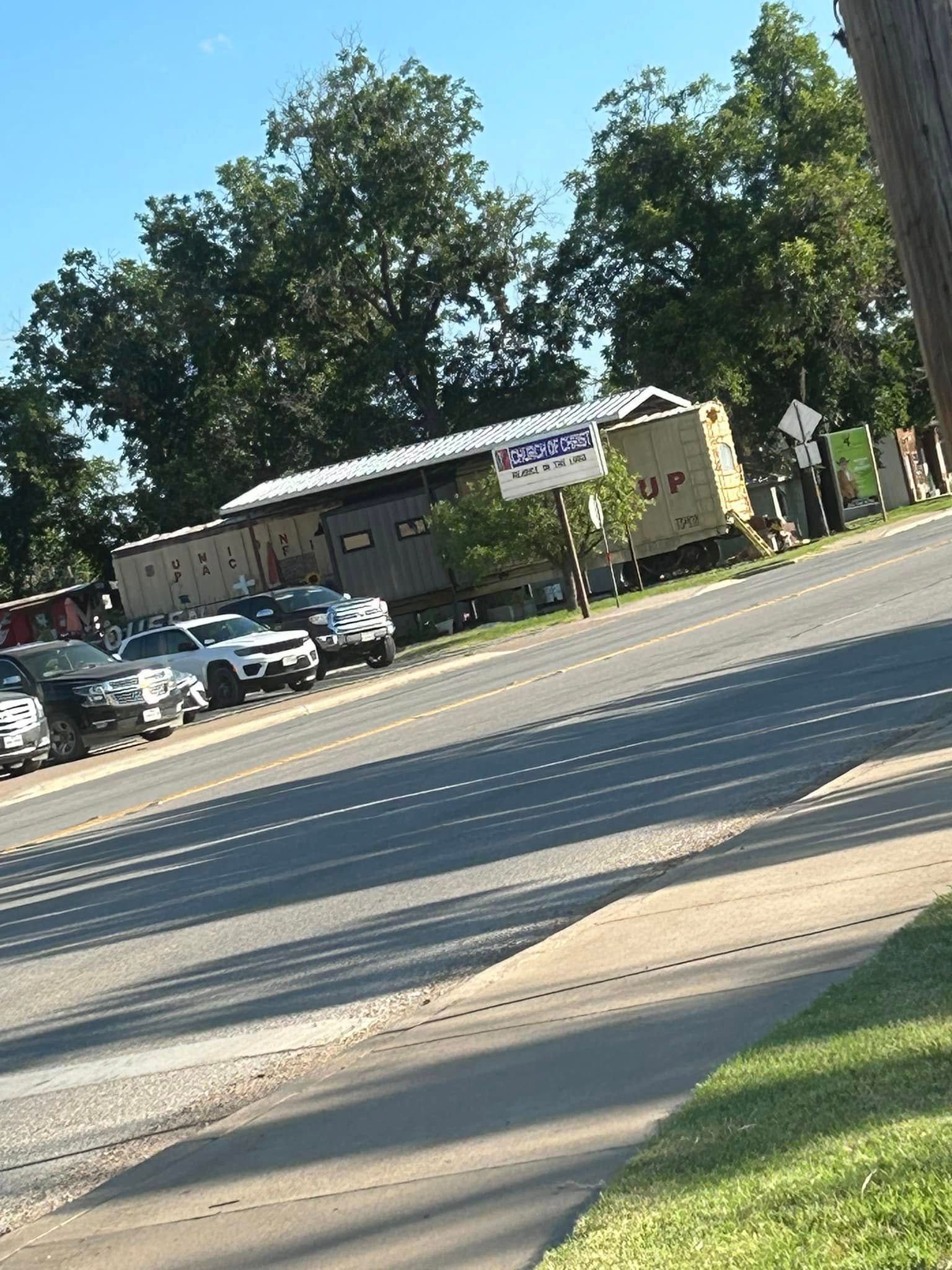 A tan railroad boxcar with a logo sits parked next to a paved road with several cars parked in a row.