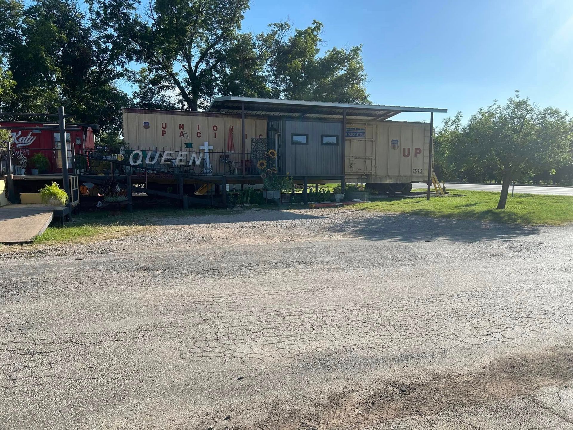 An old train caboose used as a structure, positioned on a gravel lot next to a red shipping container under trees.