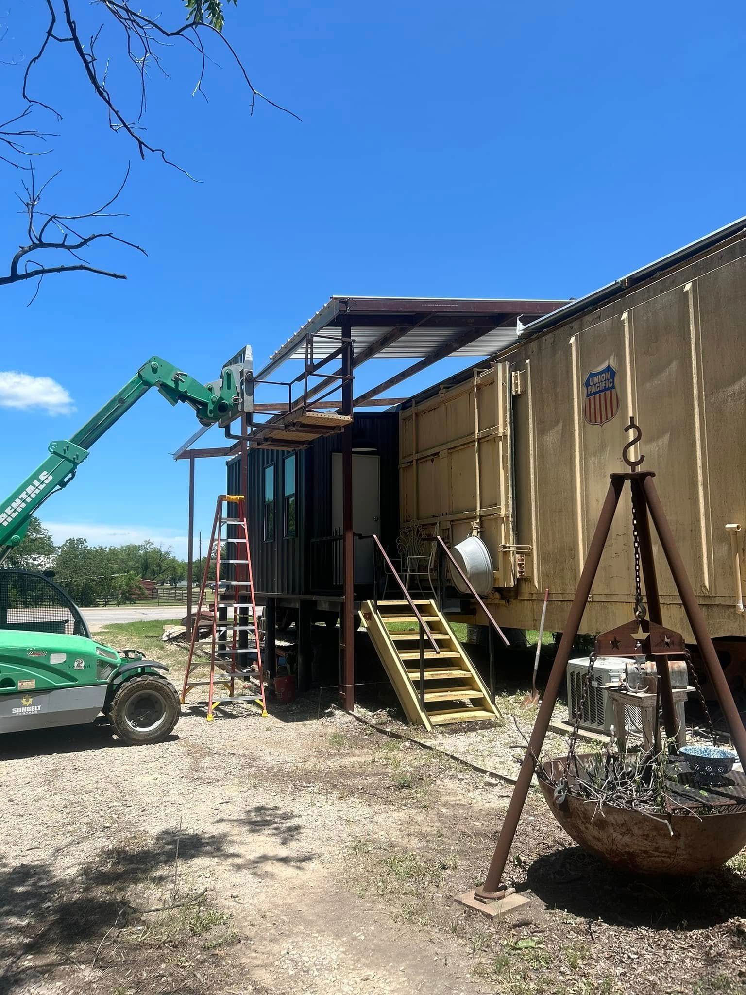 A yellow train car with an attached metal awning structure and a yellow staircase, set in a gravel area with a lift.