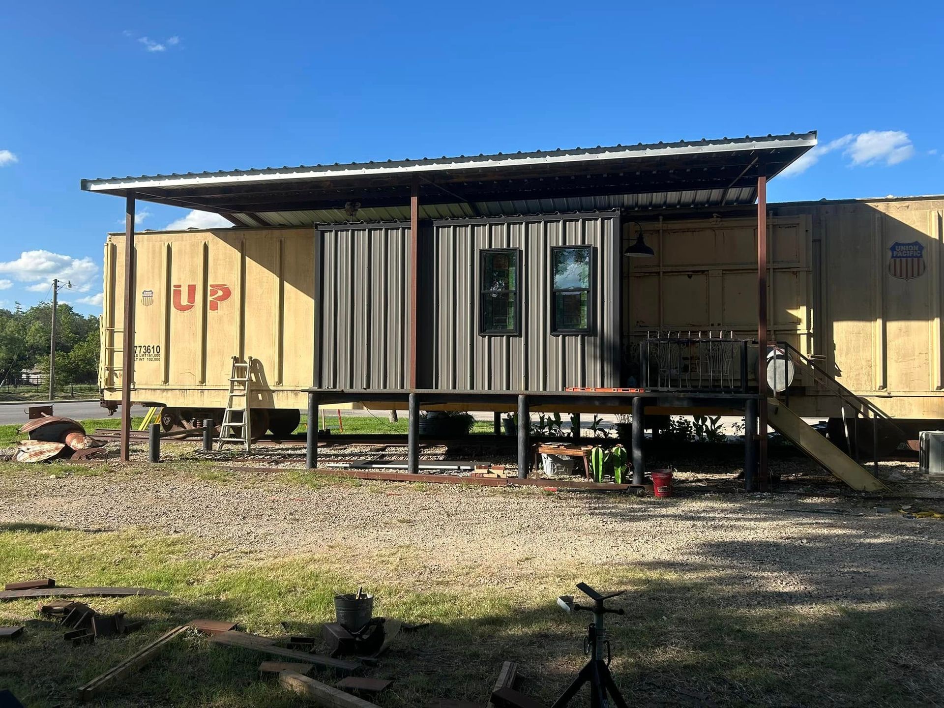 A renovated train boxcar with a modern metal entryway, porch, and roof extension, set on a gravel lot under a blue sky.