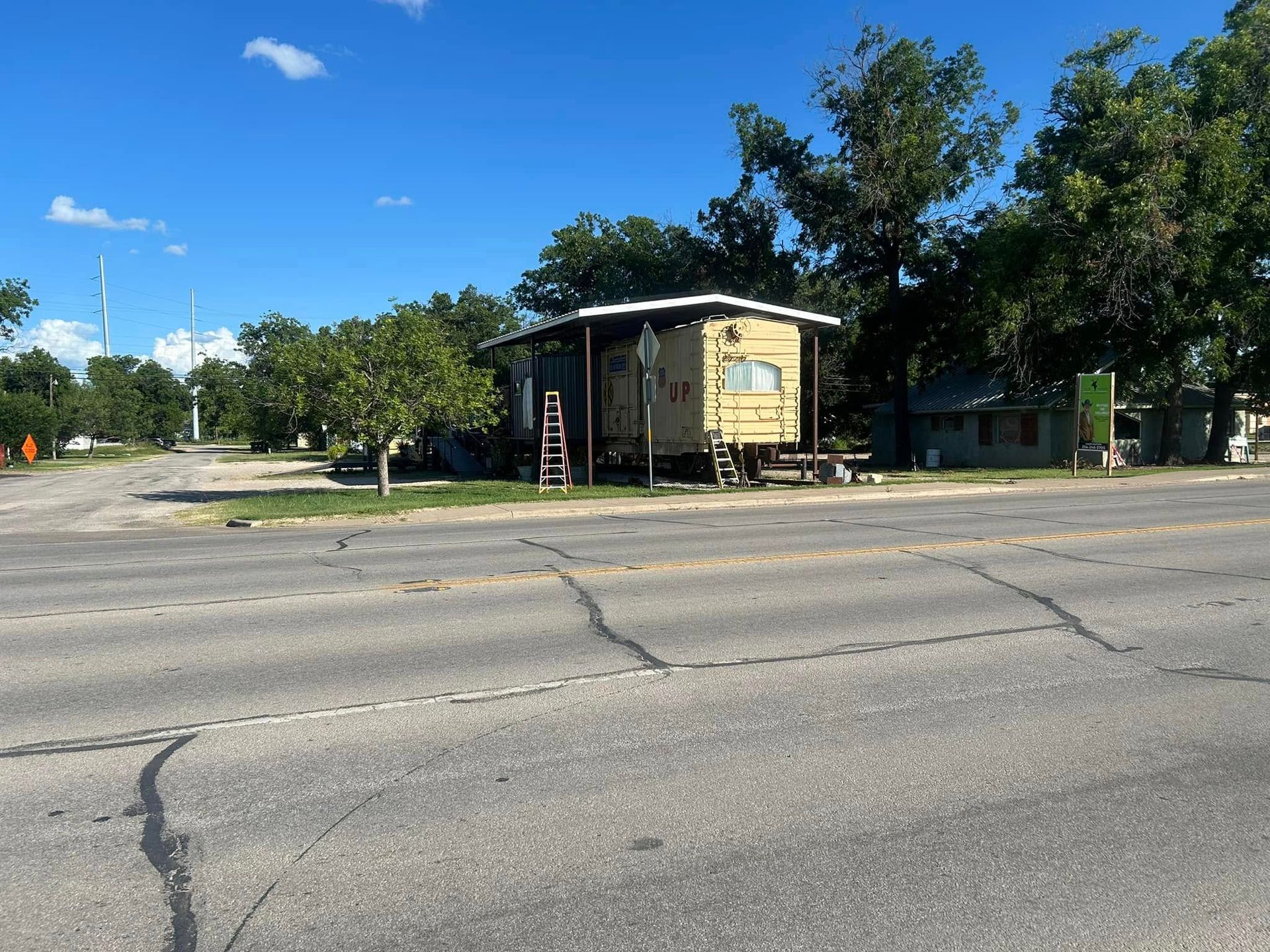 A tan manufactured home elevated on blocks sits near a paved road under a bright blue sky with scattered trees.