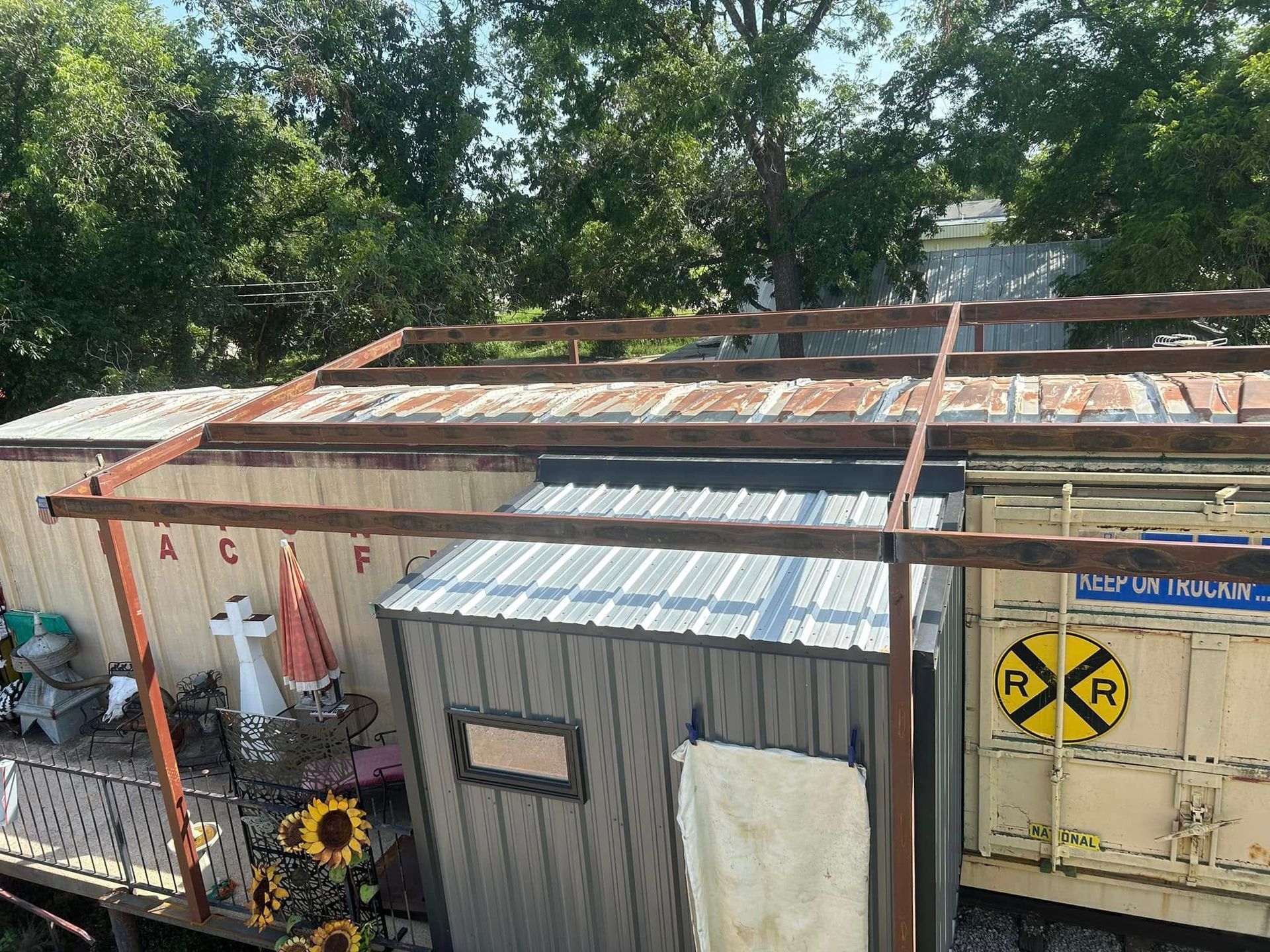 A metal frame structure stands over an outdoor area with a garden shed and a railroad car featuring a crossing sign.
