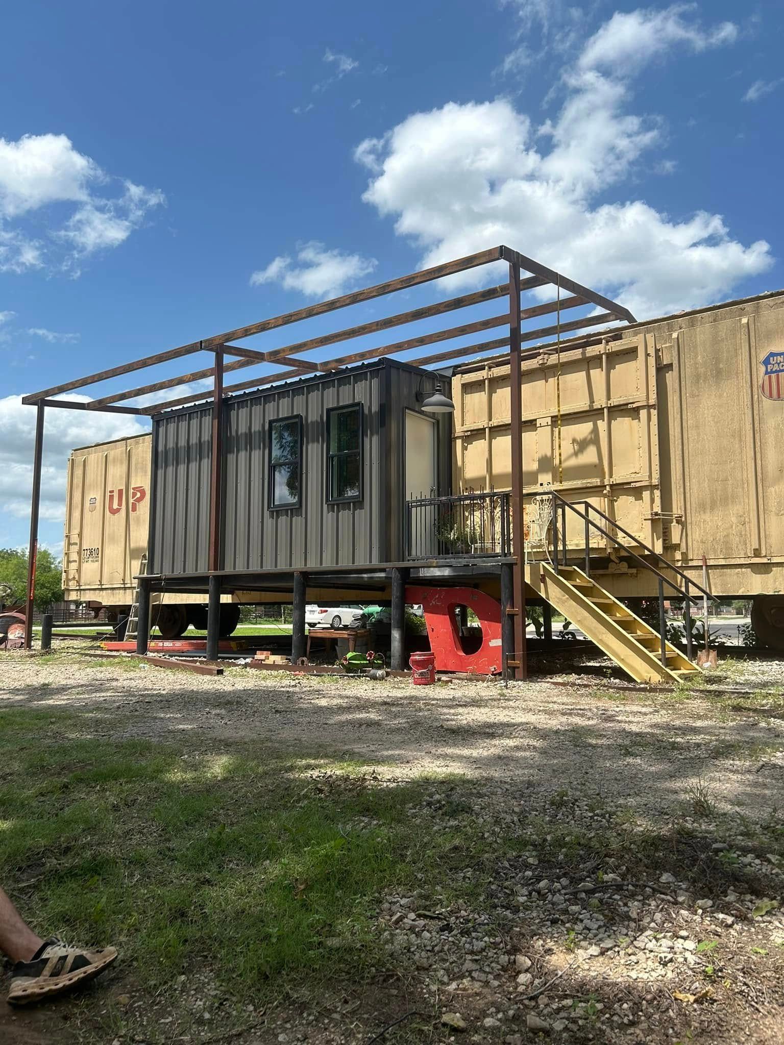 A dark shipping container structure with a metal frame sits between two train cars under a bright, cloudy sky.