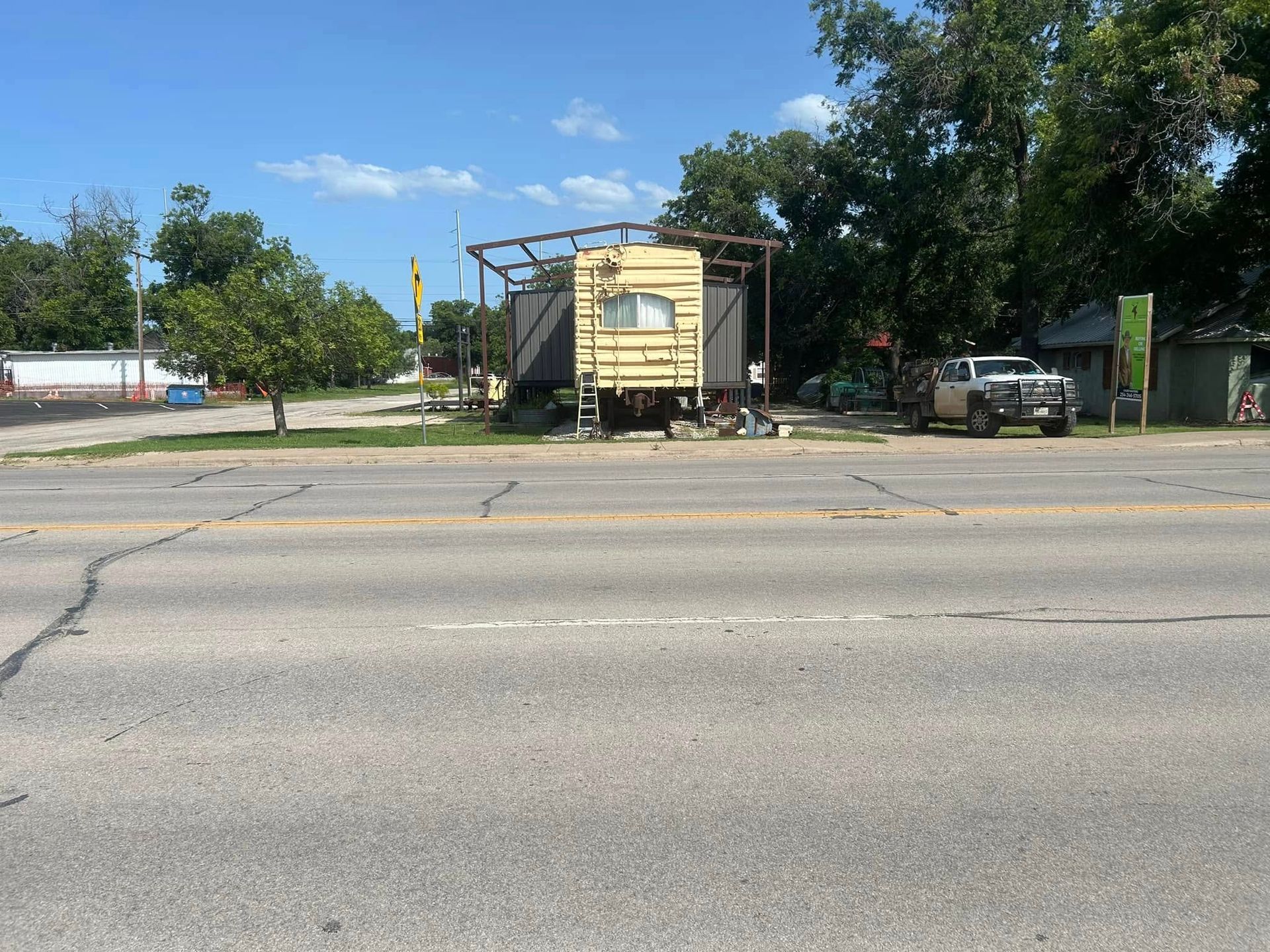 A yellow, weathered mobile home sits behind a metal frame structure next to a pickup truck along a paved road.