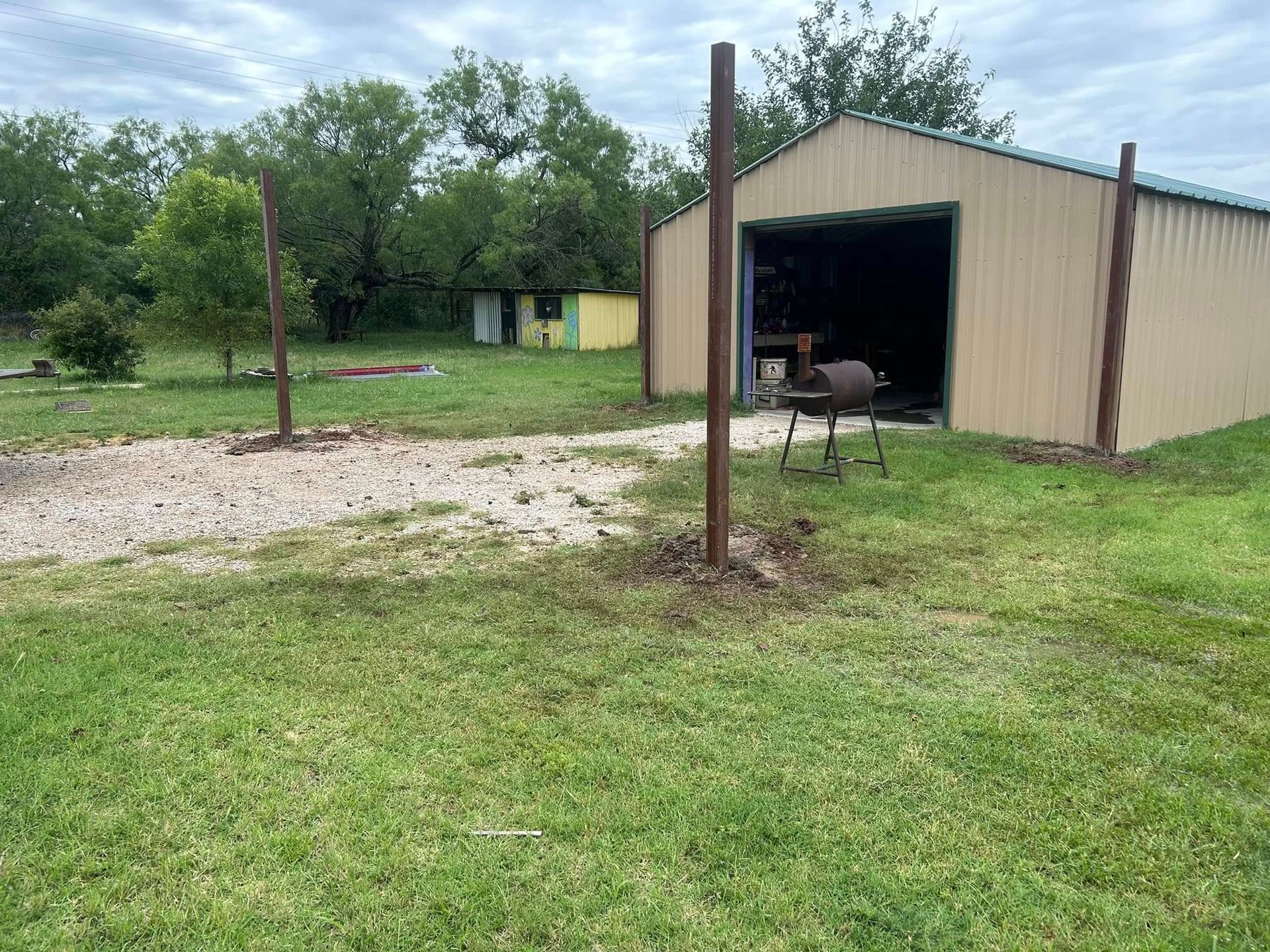 A grassy yard with a metal garage, a small shed in the distance, and two upright metal posts in the foreground.