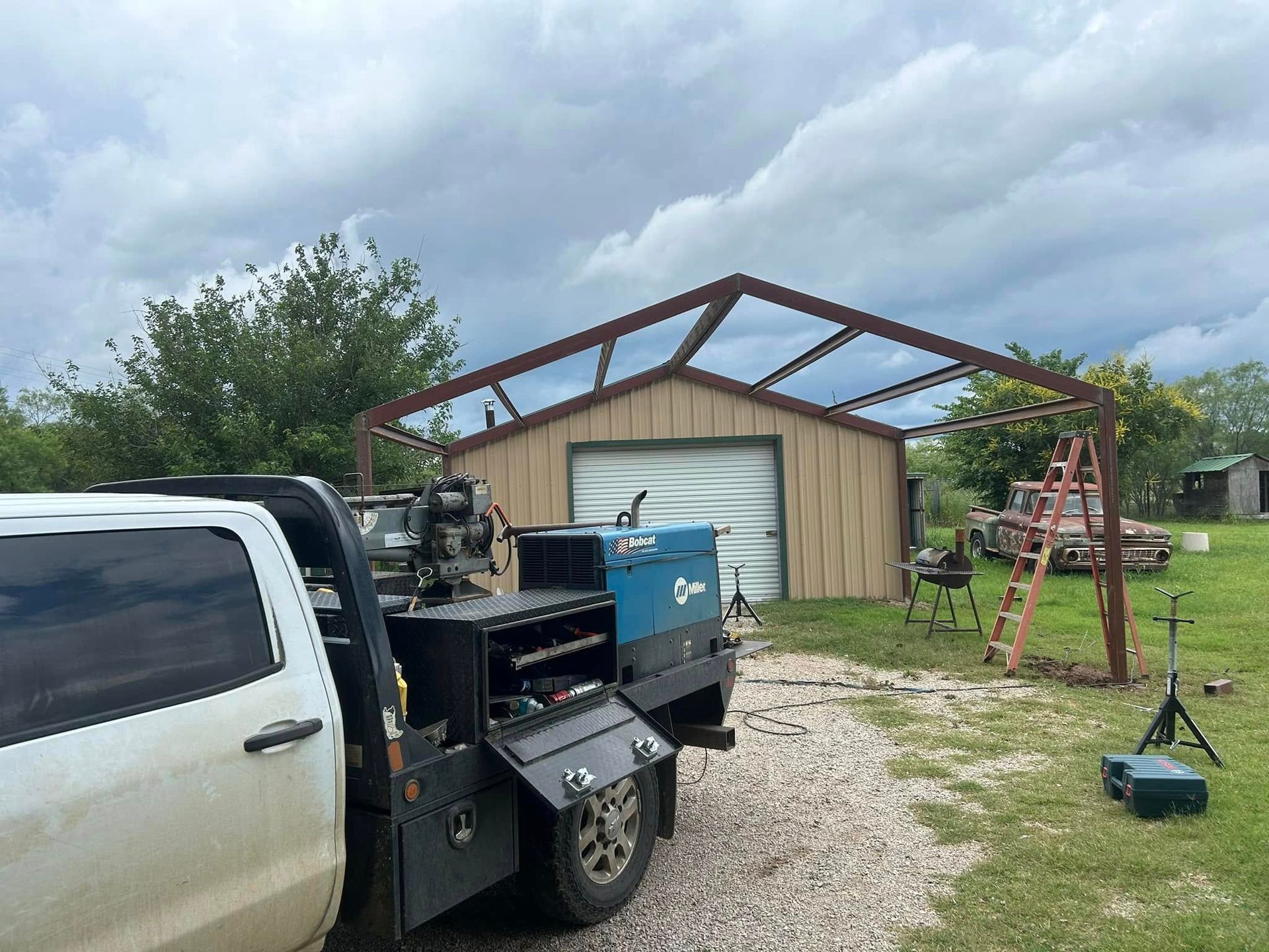 A work truck with a blue welding machine on its bed parked near a steel barn frame under construction on a grassy lot.