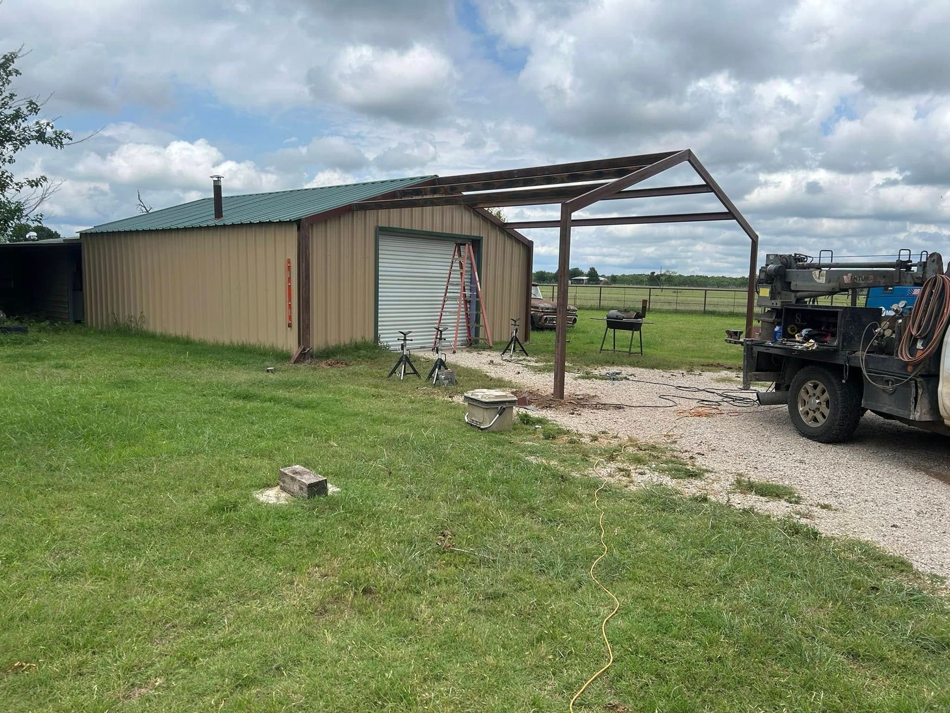 A tan metal shop building with a green roof, partially open canopy structure, and a service truck parked on a gravel drive.