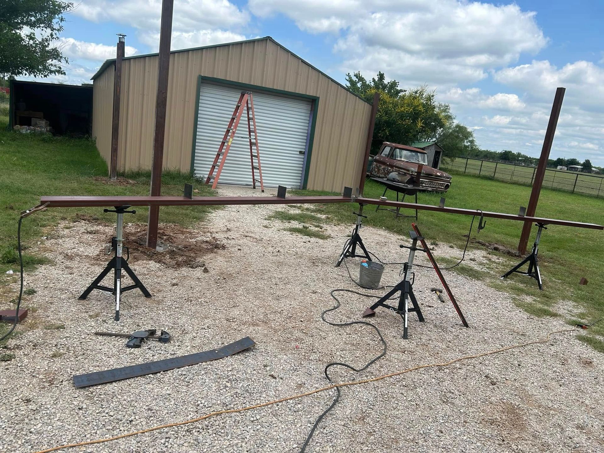 A metal rail supported by jack stands in a gravel lot, with a garage, ladder, and field in the background.