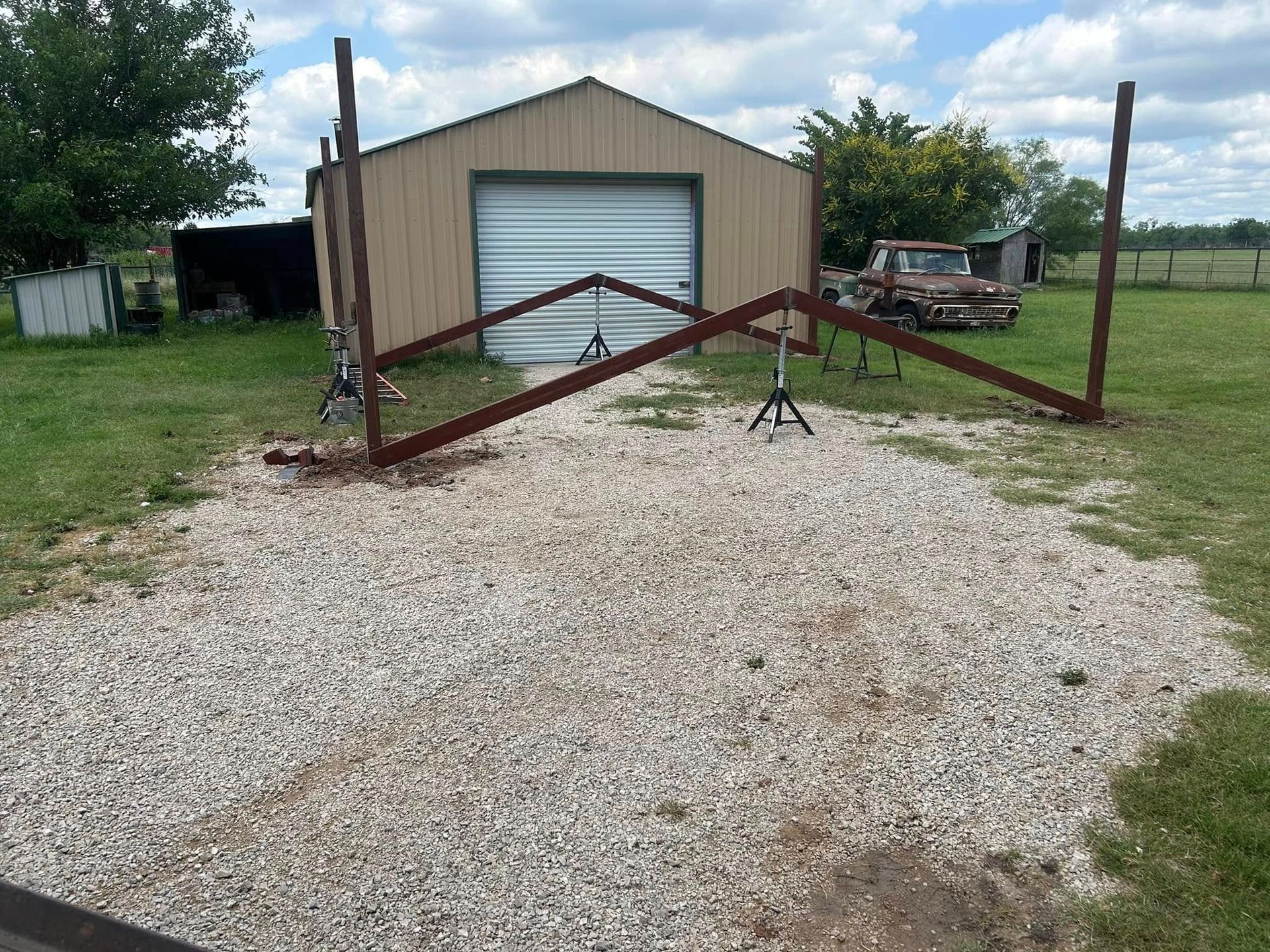 A metal gate frame structure stands on a gravel driveway in front of a tan metal building with a truck parked nearby.