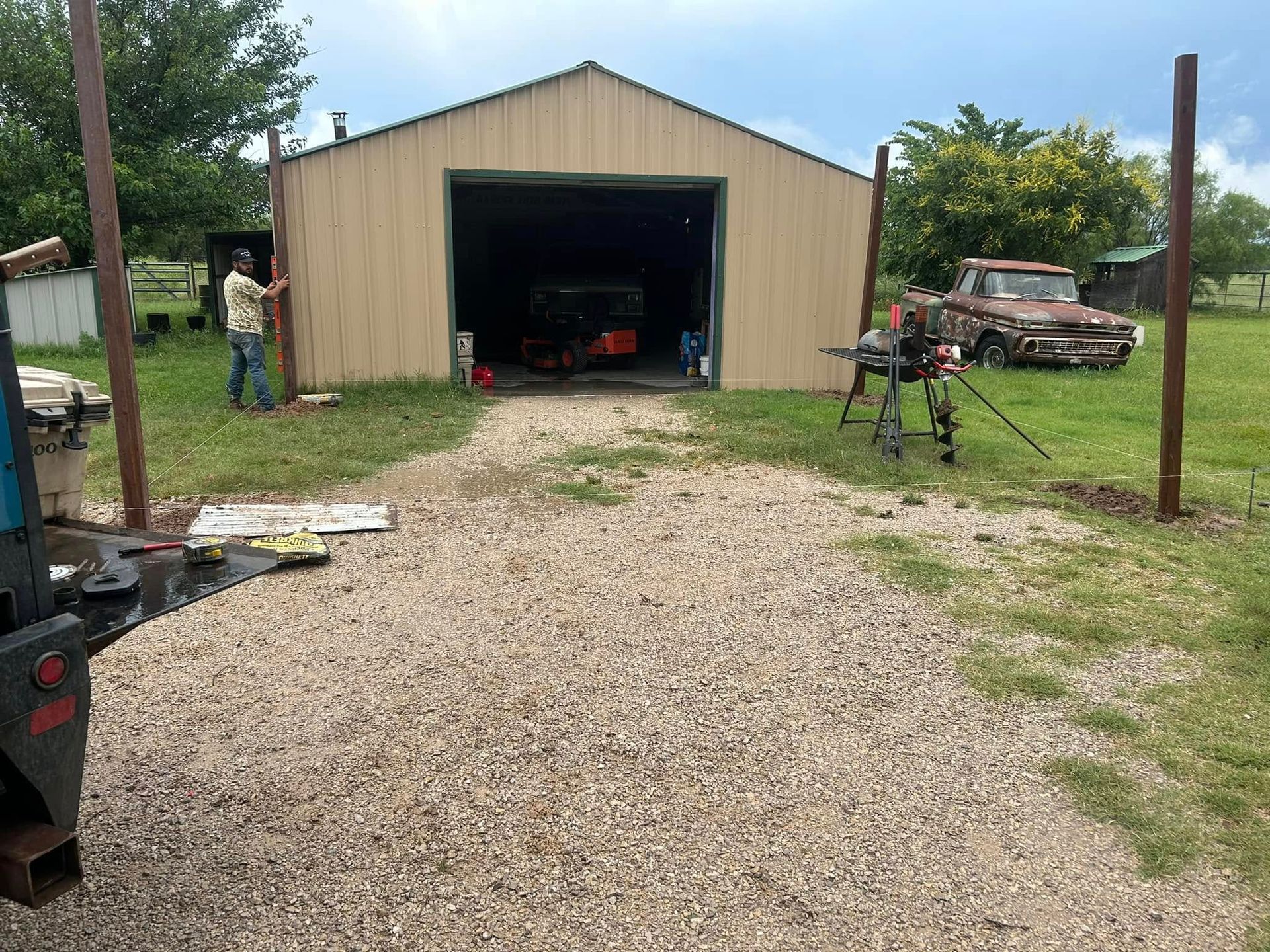 A man stands beside a metal storage building with an open garage door, set in a rural area with an old truck nearby.