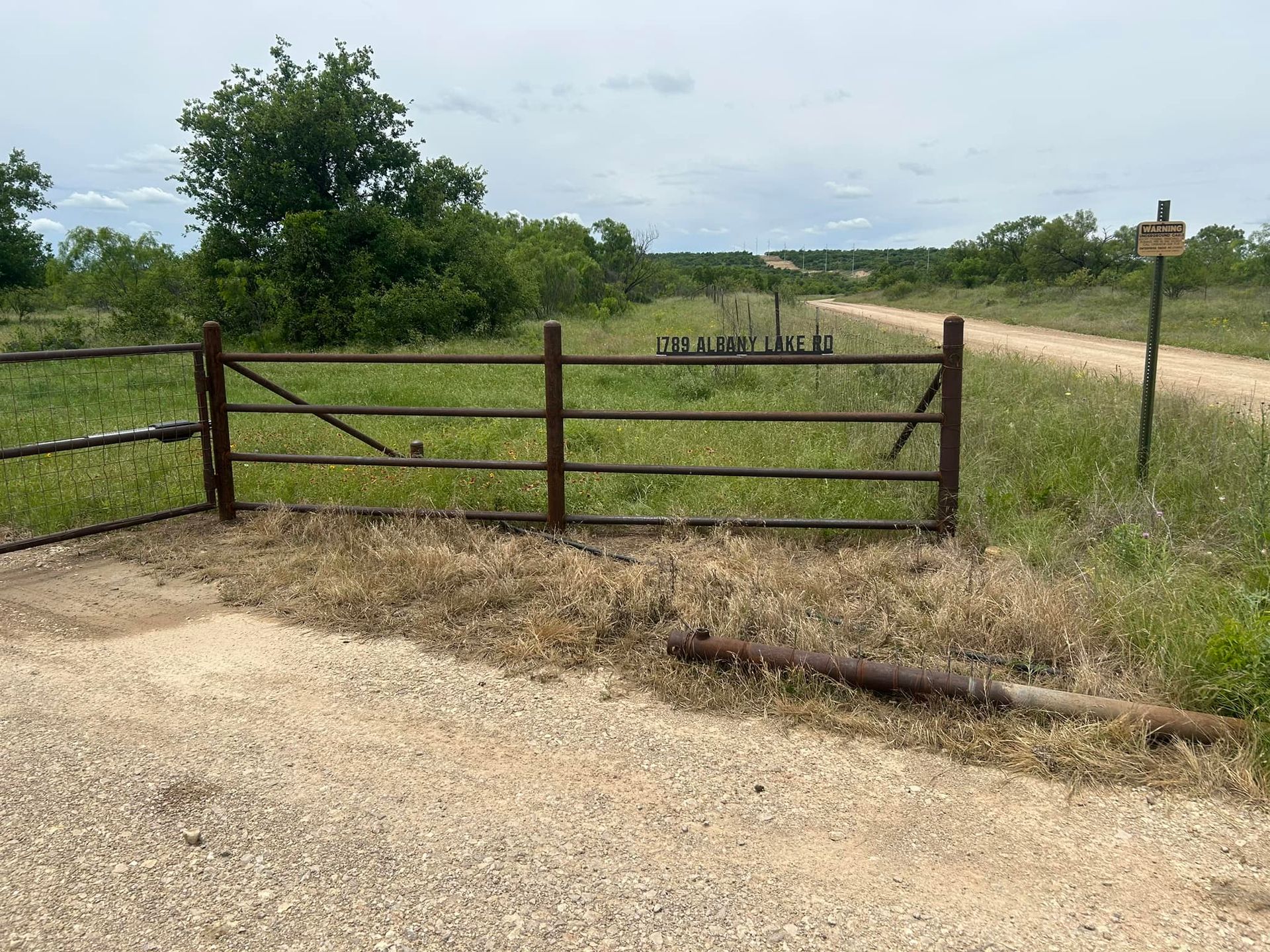 A metal gate stands at the entrance of a gravel path in a grassy field under a partly cloudy sky.