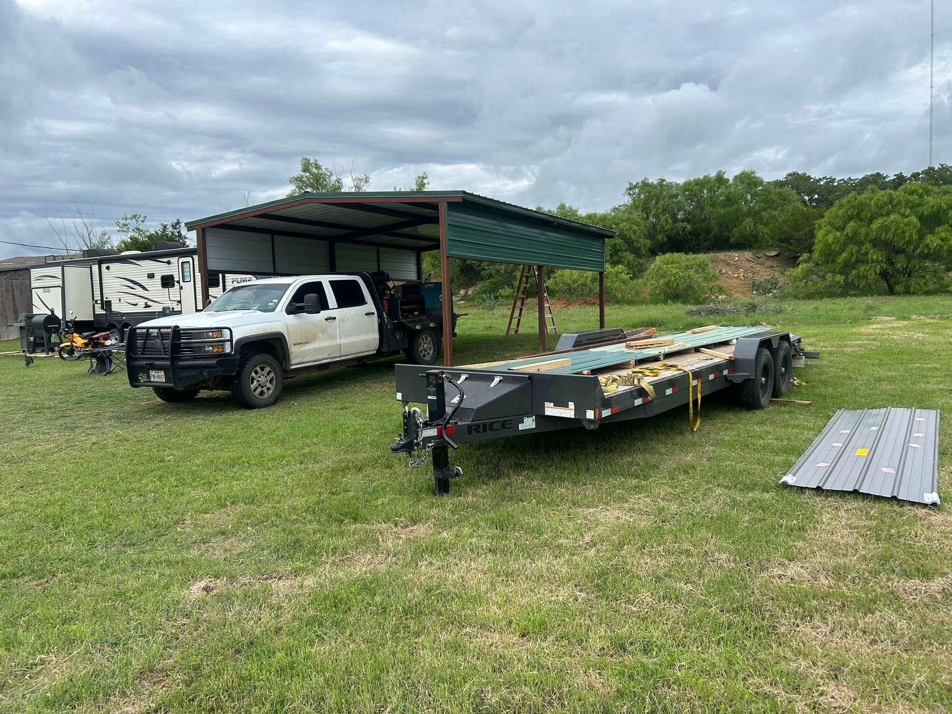 A white truck parked under a metal carport next to a trailer in a grassy field, with a piece of metal siding nearby.