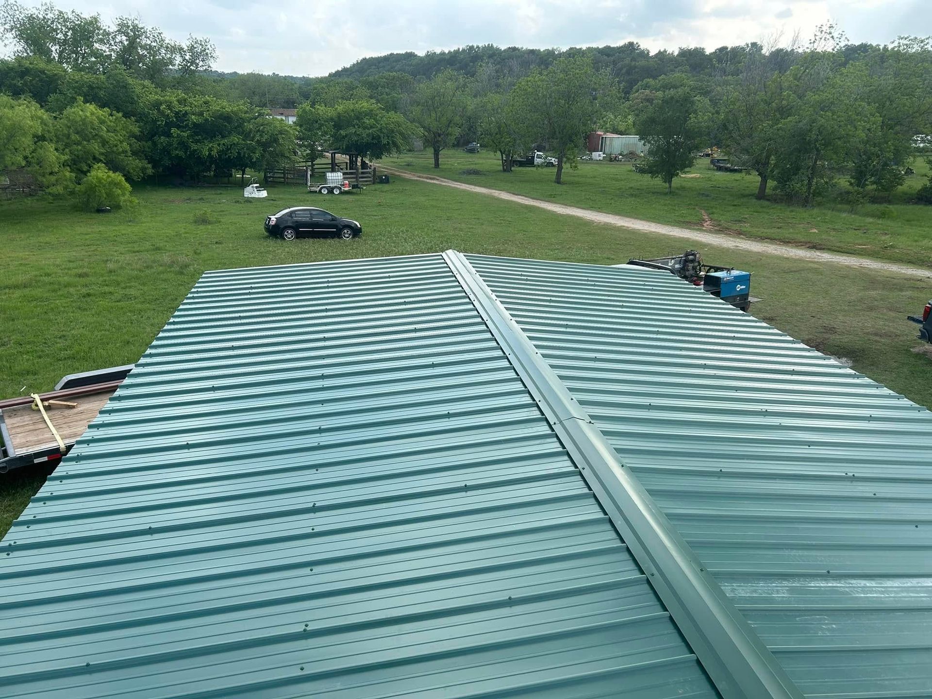 A high-angle view of a green, rippled metal roof on a building, overlooking a rural landscape with trees and a car.